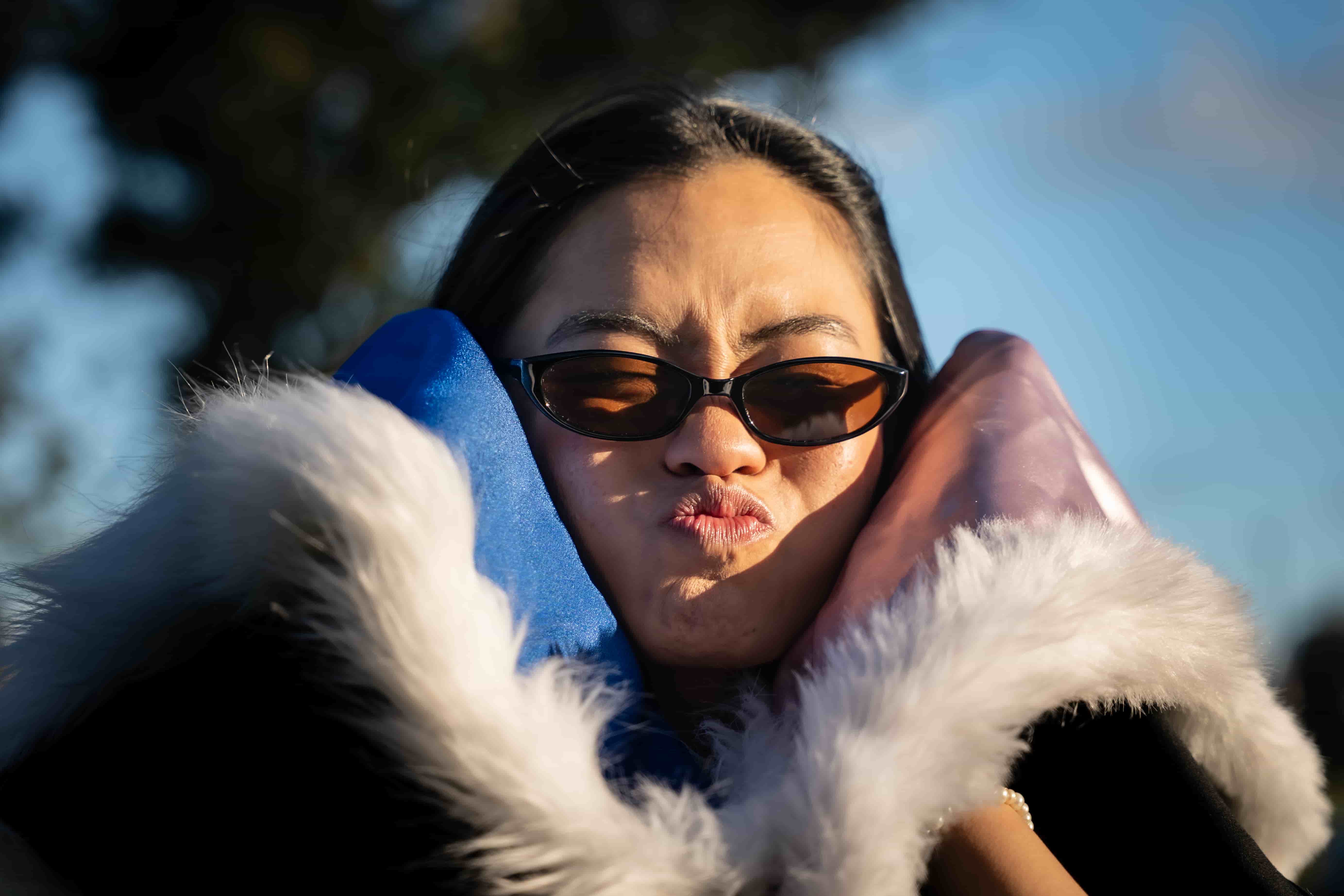 Ruby in sunglasses pulling a face, wrapped in the blue and pink fur-trimmed hood of her graduation gown