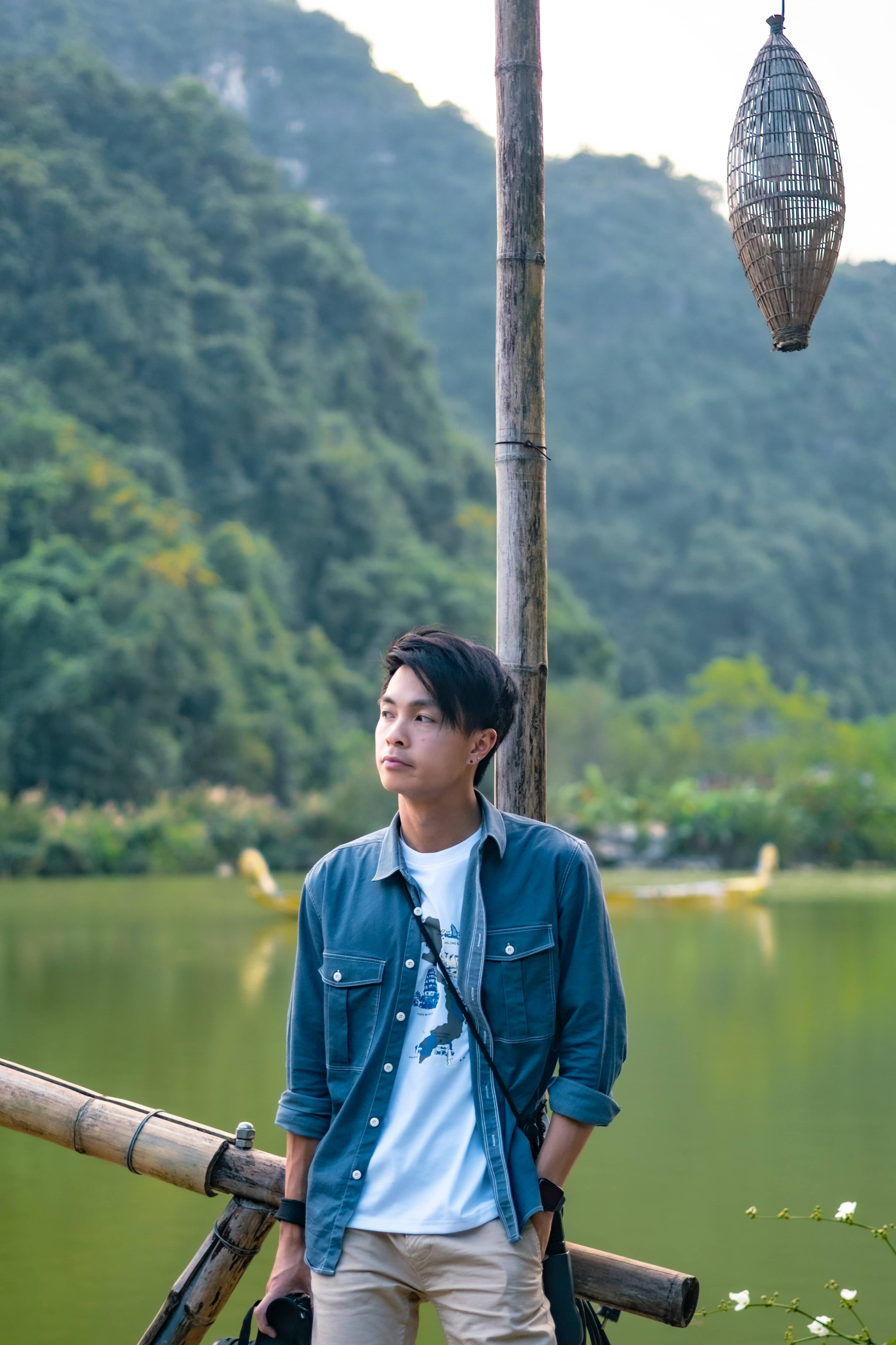 Man in a denim jacket by a bamboo rail at a green lake, forested limestone hills behind, Vietnam