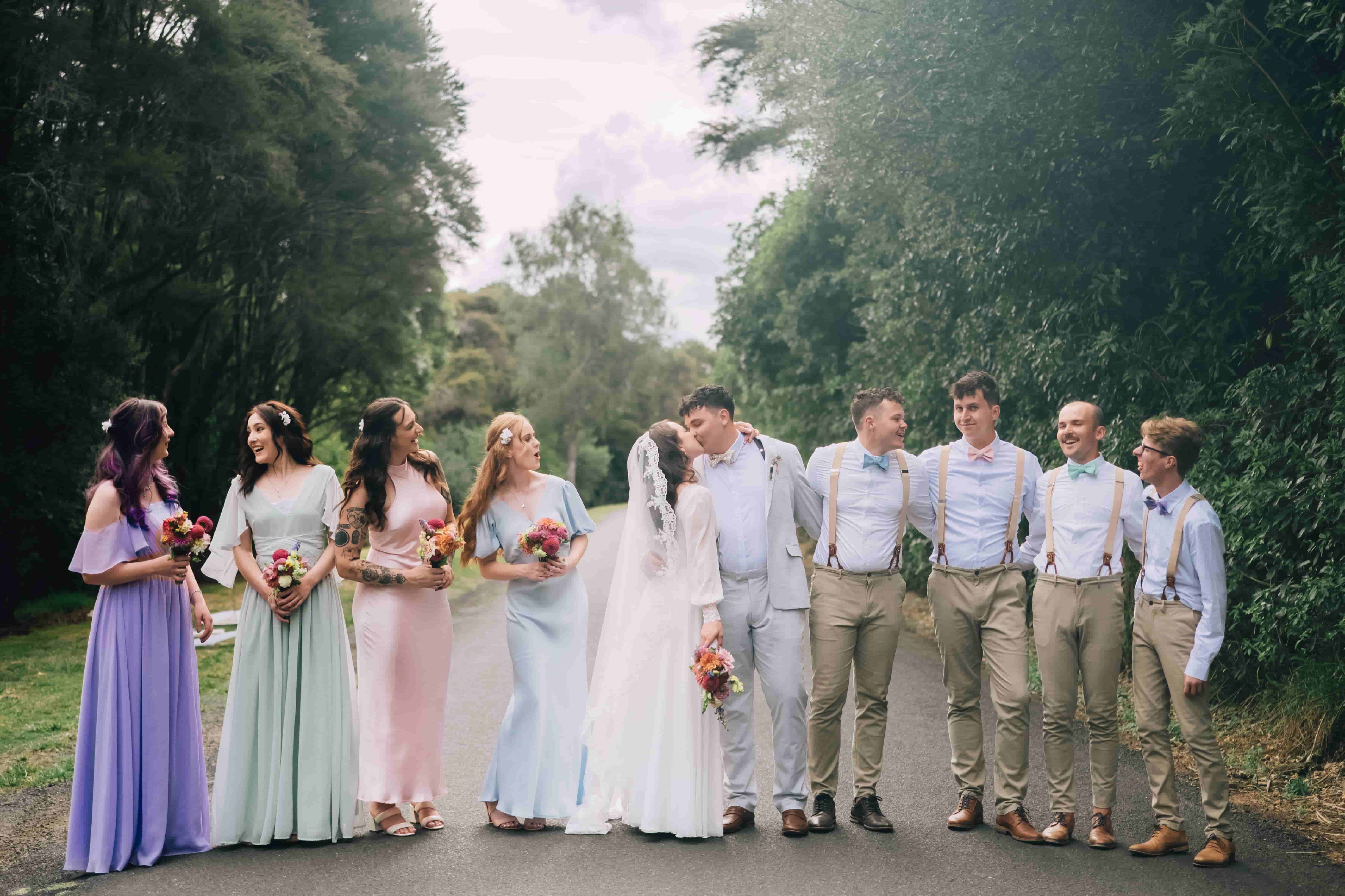Mia and Joe kissing on a tree-lined road, surrounded by bridesmaids in pastel dresses and groomsmen in braces