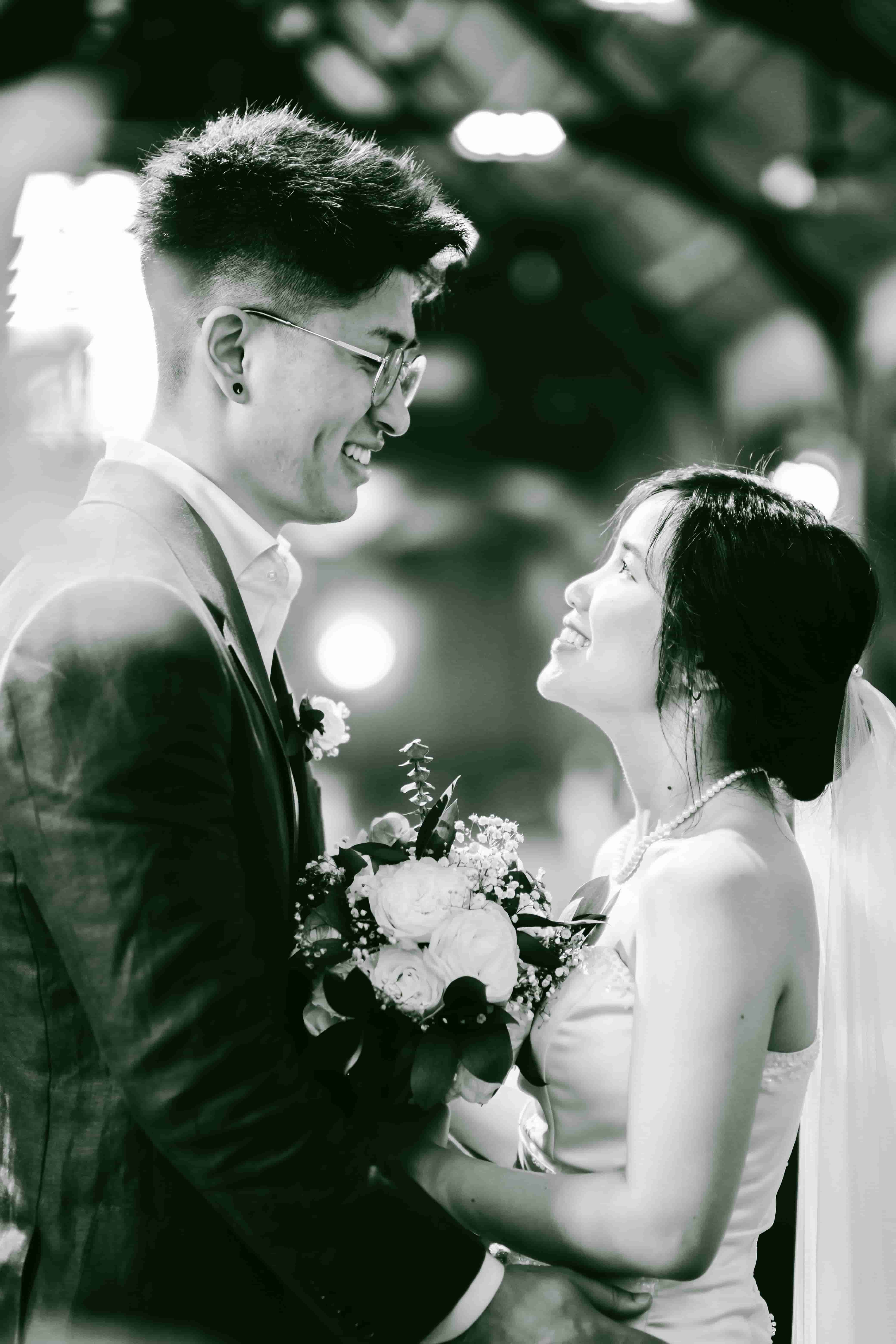 Black and white portrait of Sam and May smiling at each other, her bouquet held between them