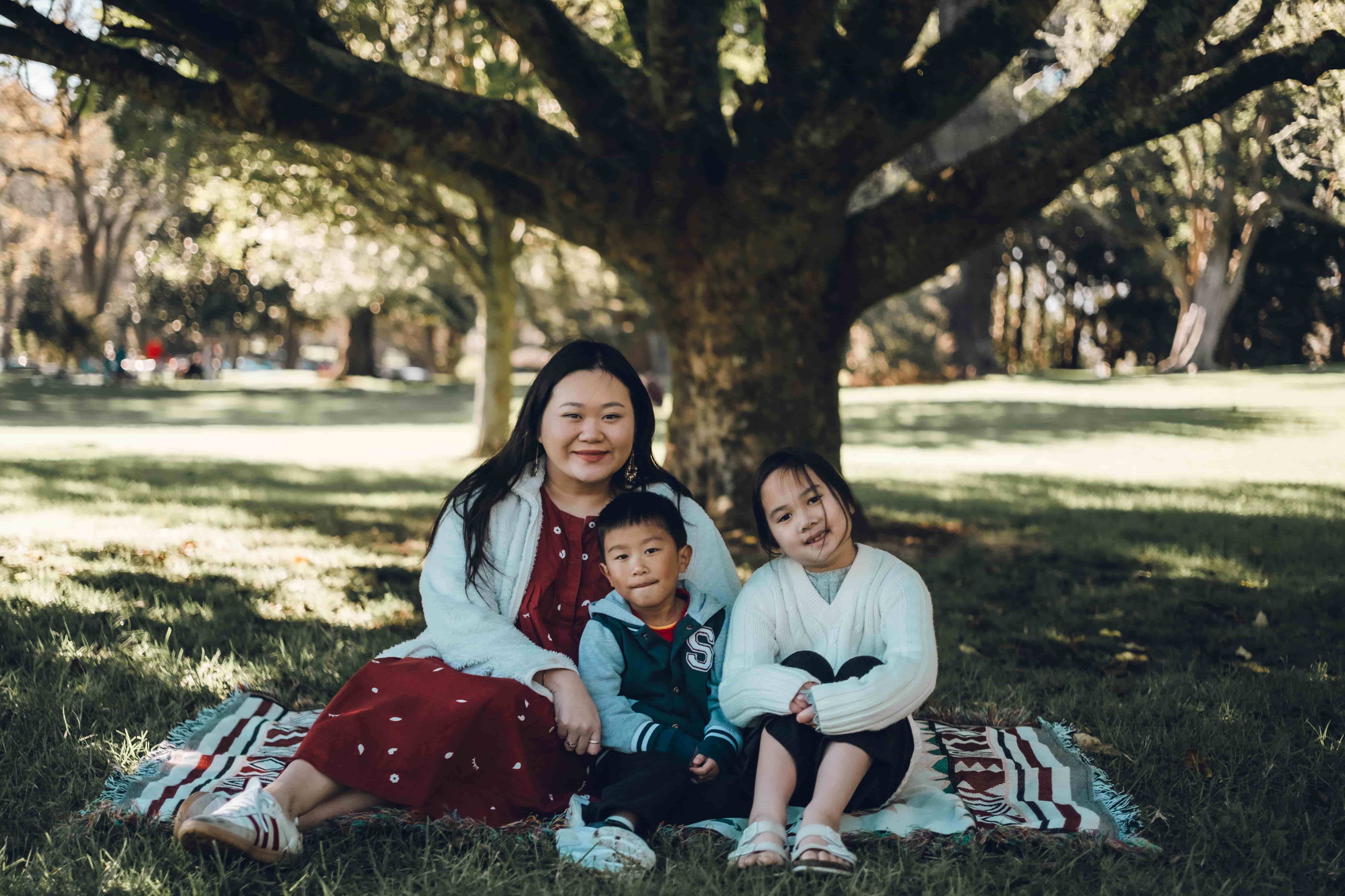 Mother sitting on a picnic blanket with two children under a wide tree