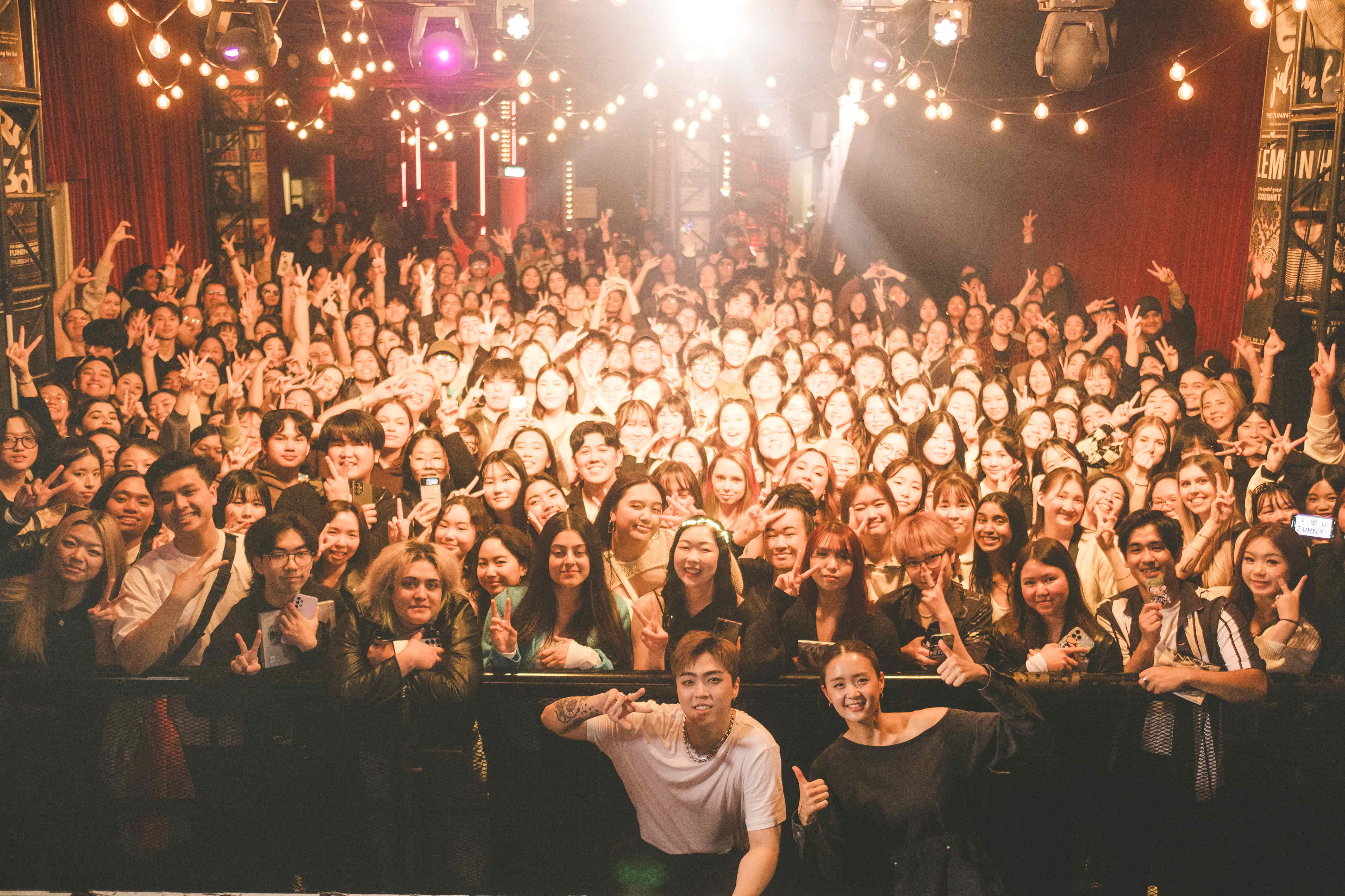Junny posing for a group photo with a packed crowd of fans under strings of festoon lights