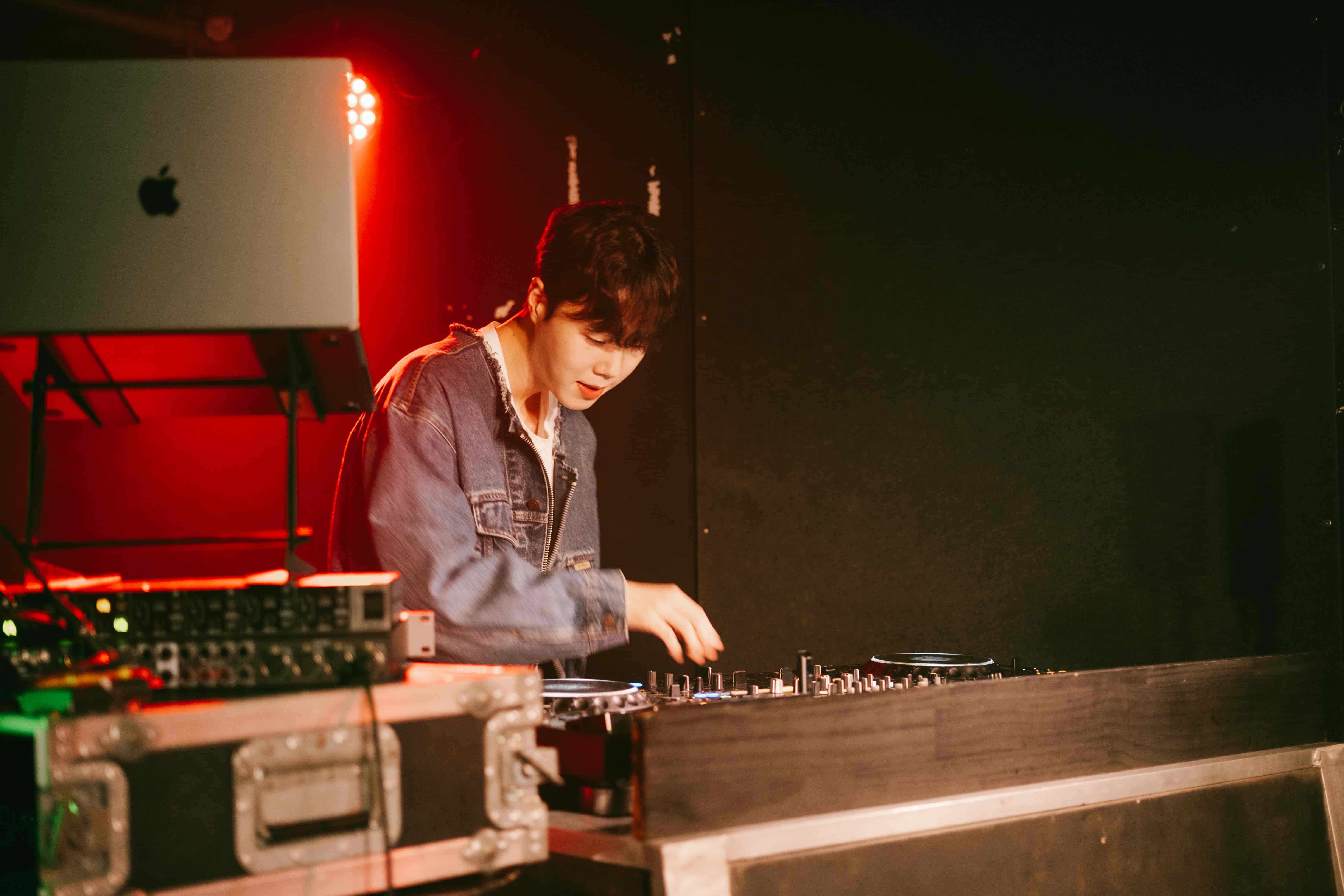 DJ leaning over the decks in a denim jacket, laptop on a stand and a red wash of light behind