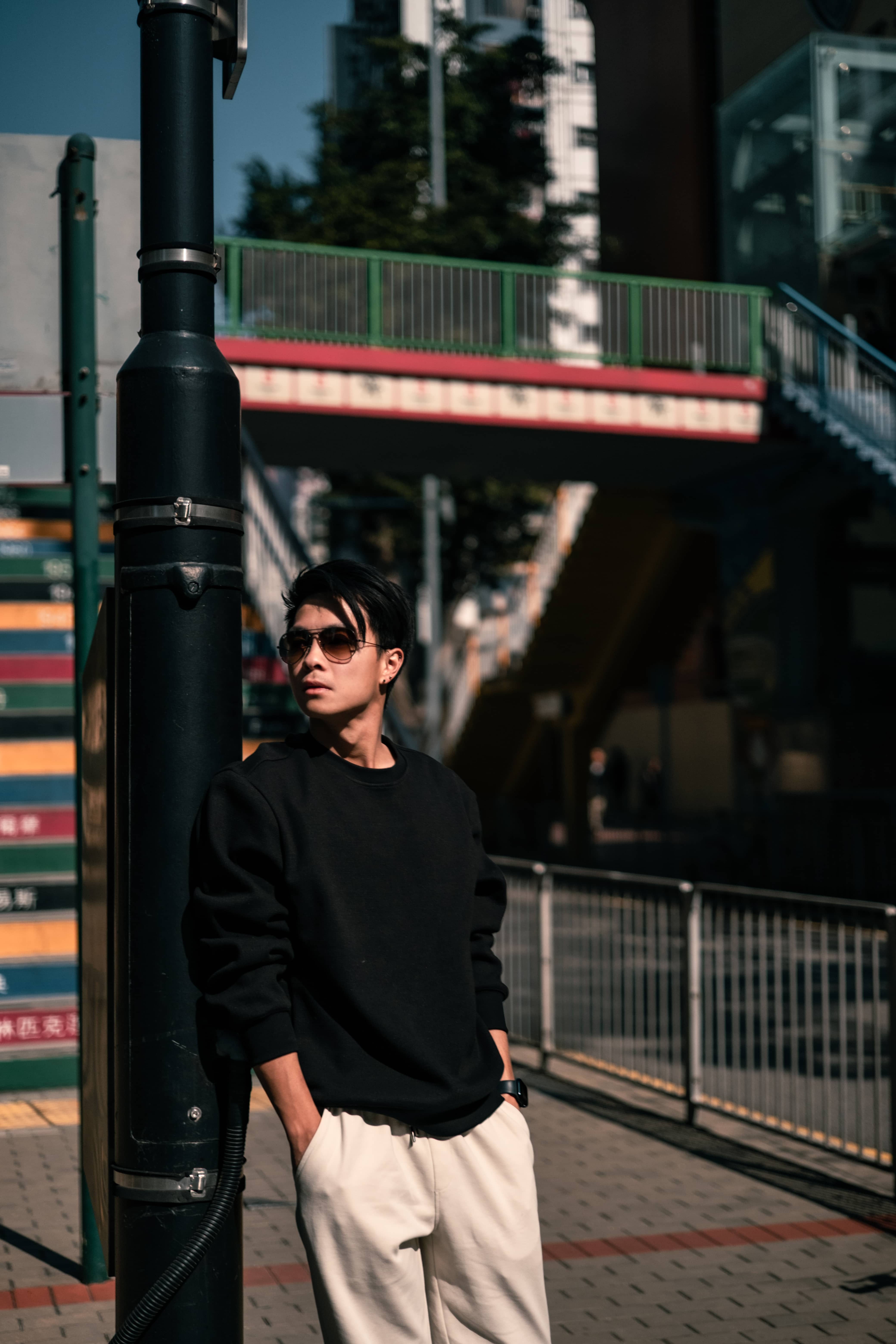 Man in sunglasses and a black sweater leaning on a lamp post by a Hong Kong footbridge in afternoon light
