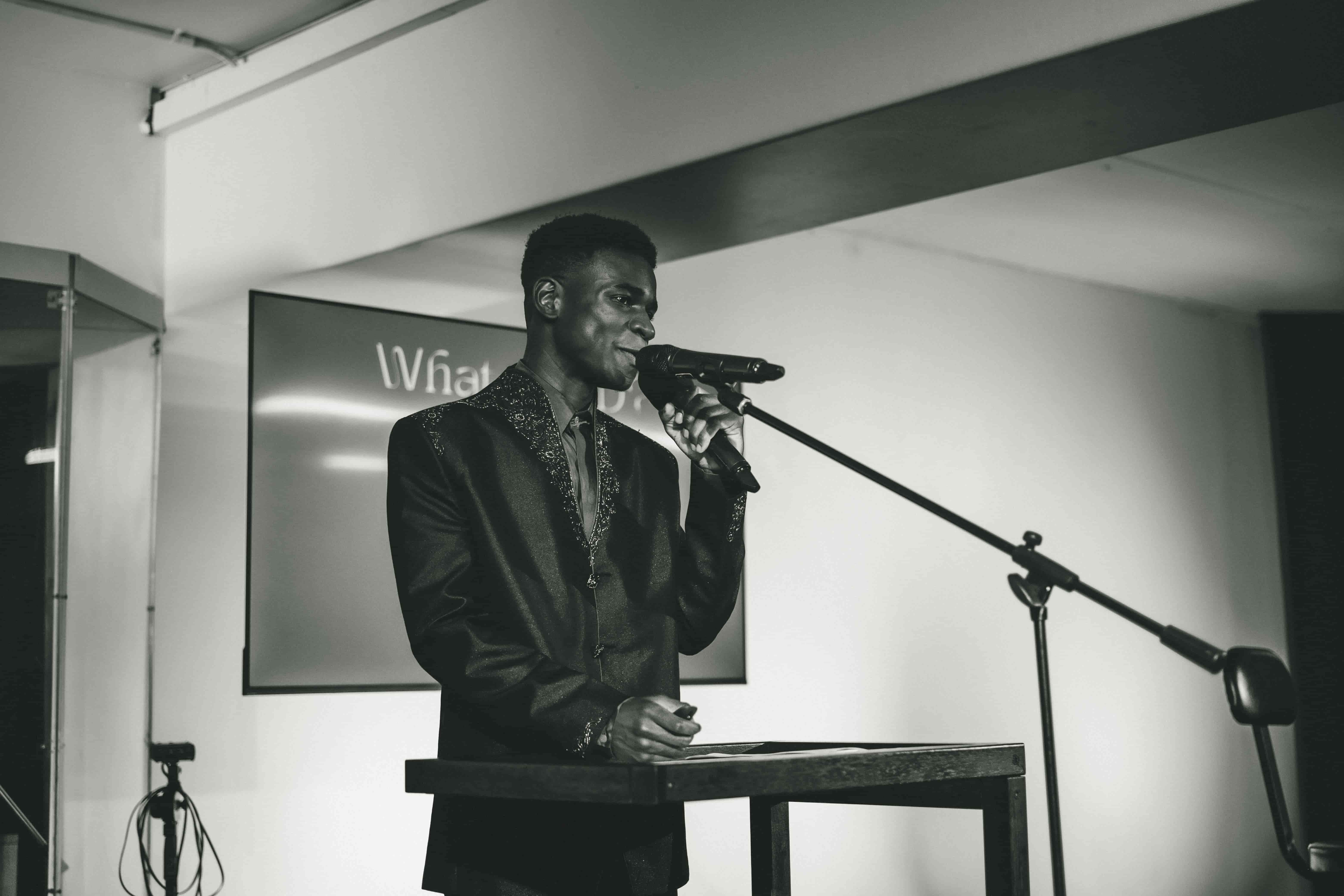 Josh in a patterned jacket at a lectern with a microphone, screen behind reading What is