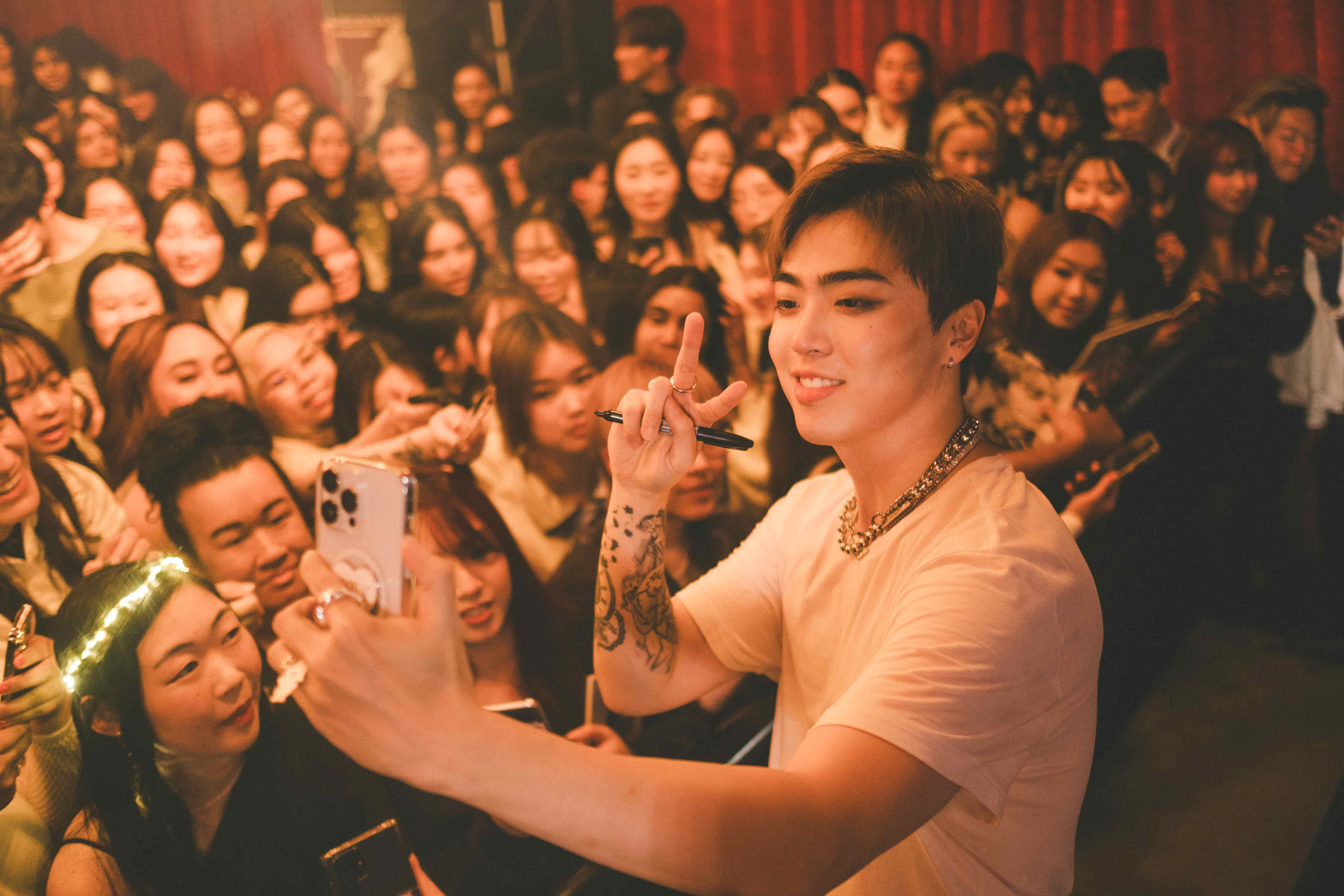 Junny holding up a phone and a pen in front of a crowd of fans against a red curtain