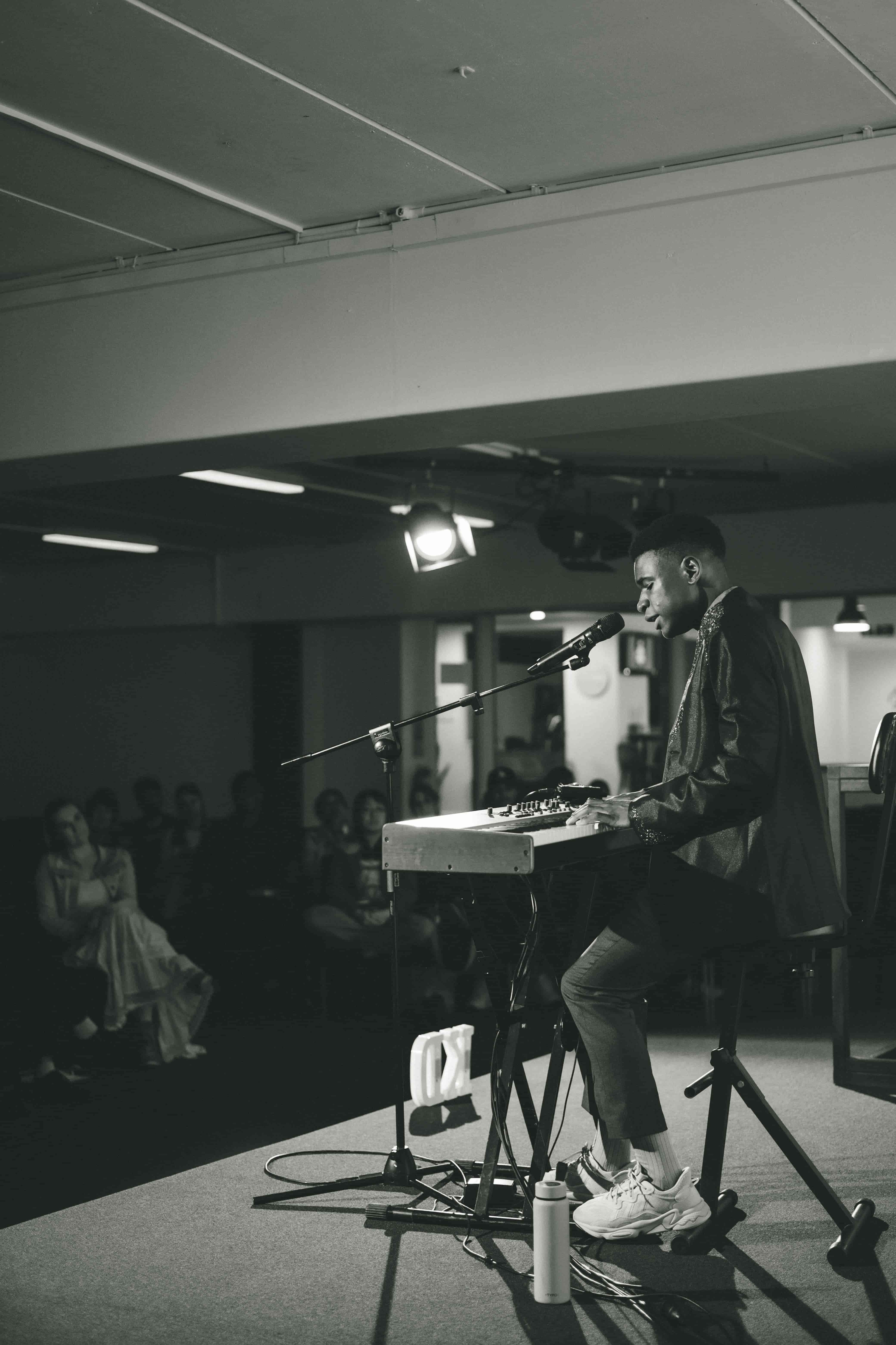 Monochrome shot of Josh singing at a keyboard on a low stage, audience faintly visible on the left