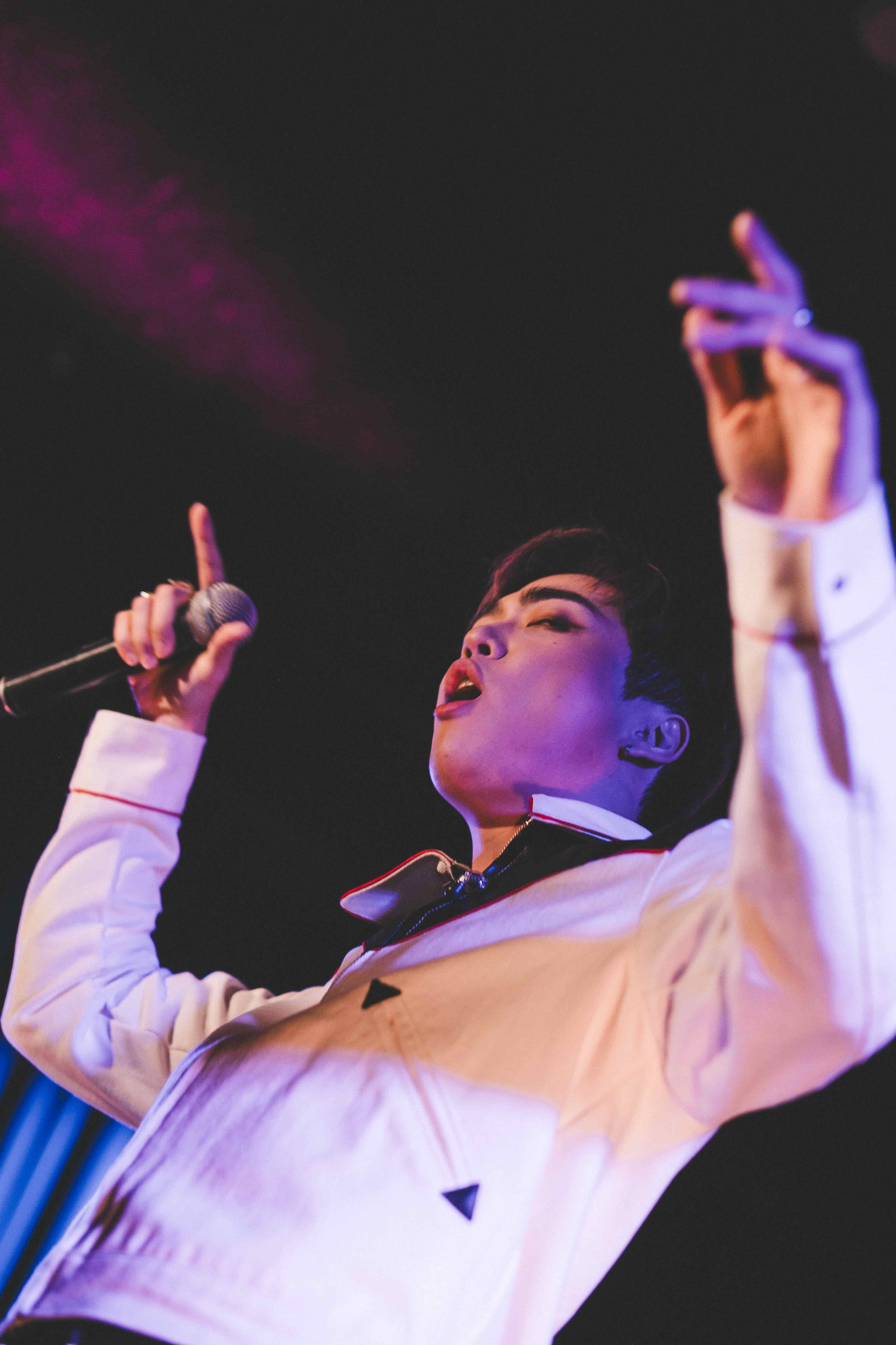Junny pointing up with both hands as he sings, shot from a low angle against a dark ceiling