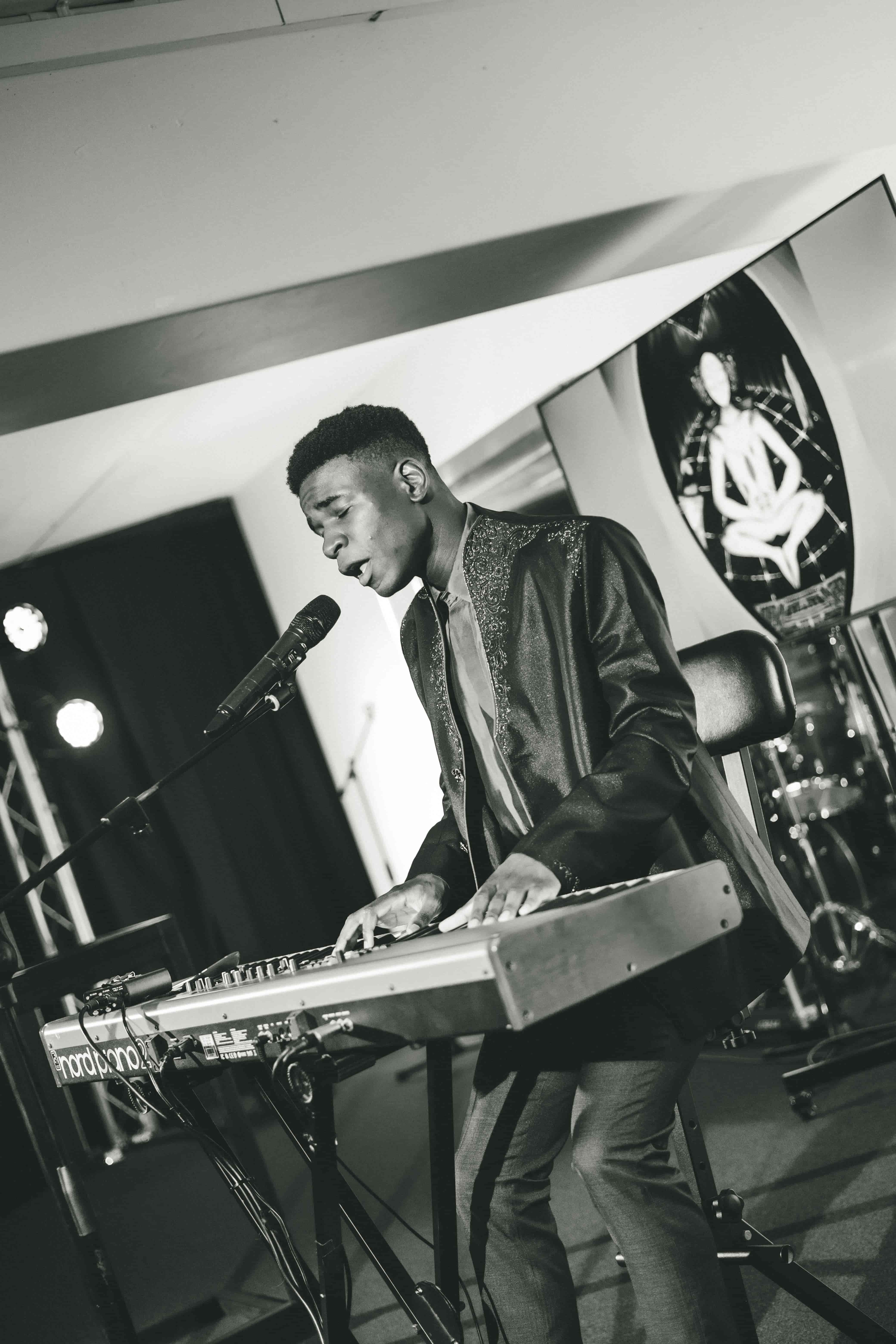 Black and white close shot of Josh singing at a keyboard, eyes lowered, stage lights behind
