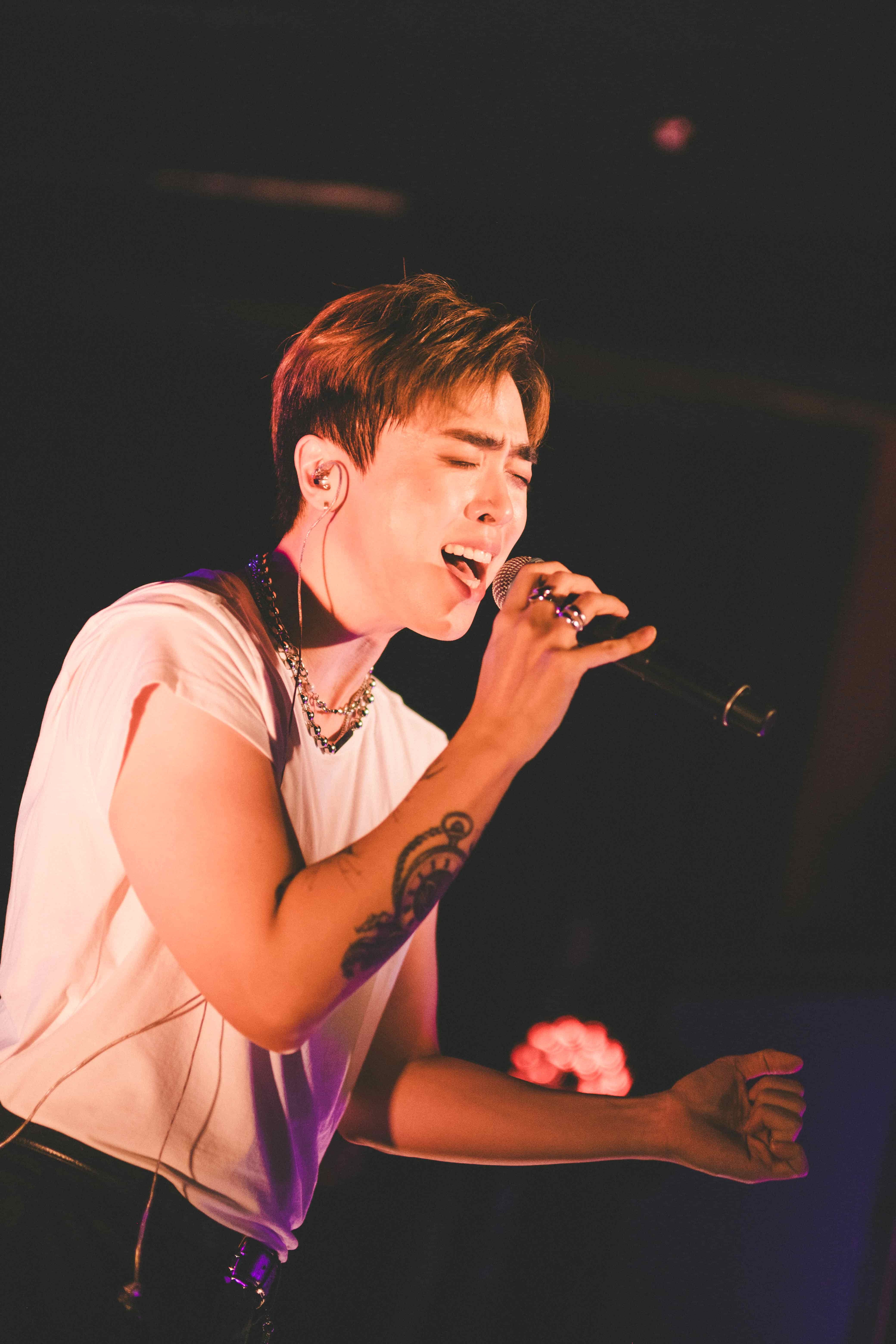 Junny in a white t-shirt singing with his arm outstretched, tattoo visible, warm light