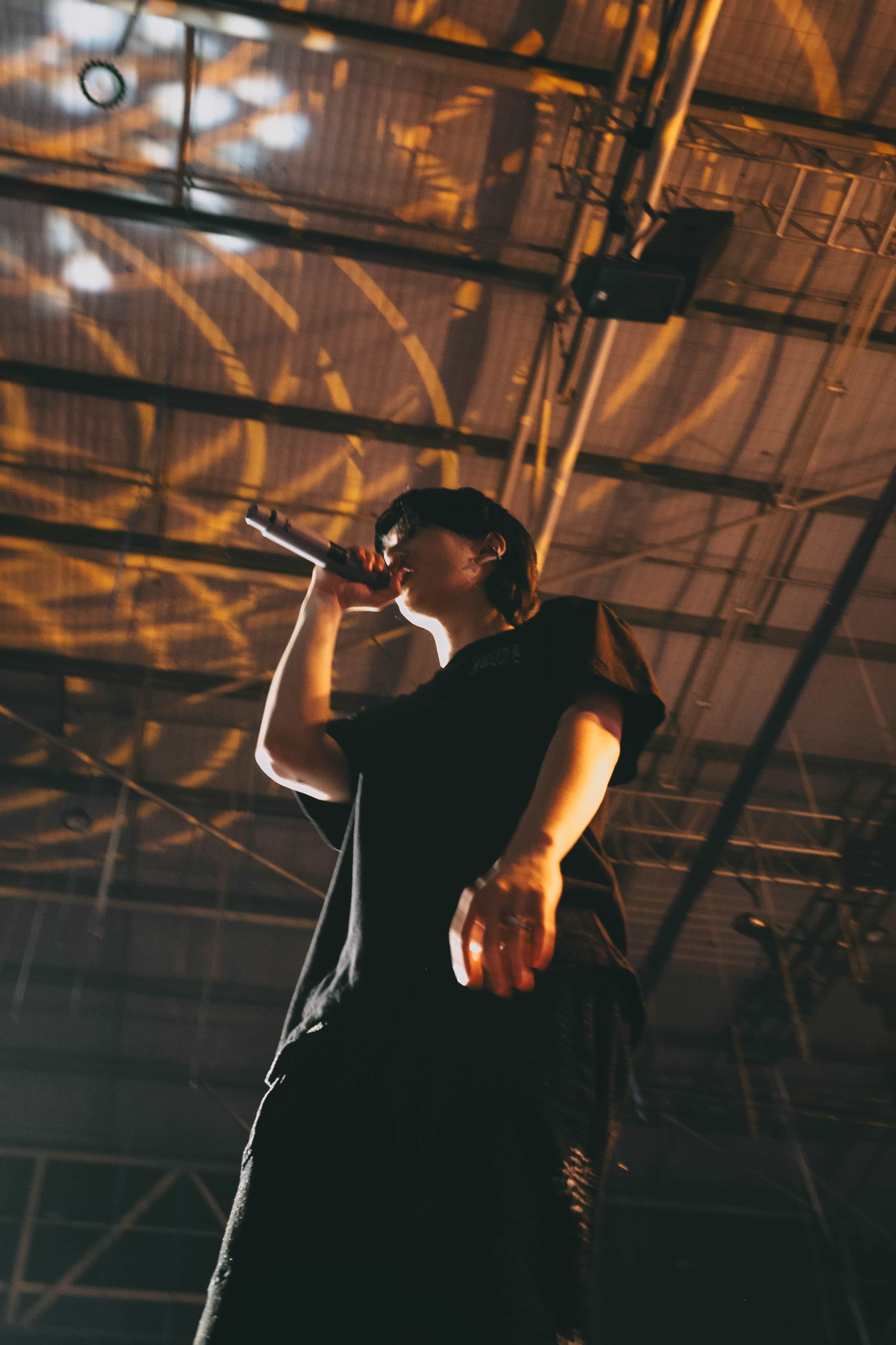 Low angle of a P1Harmony member in black singing under warm amber patterns on the arena roof