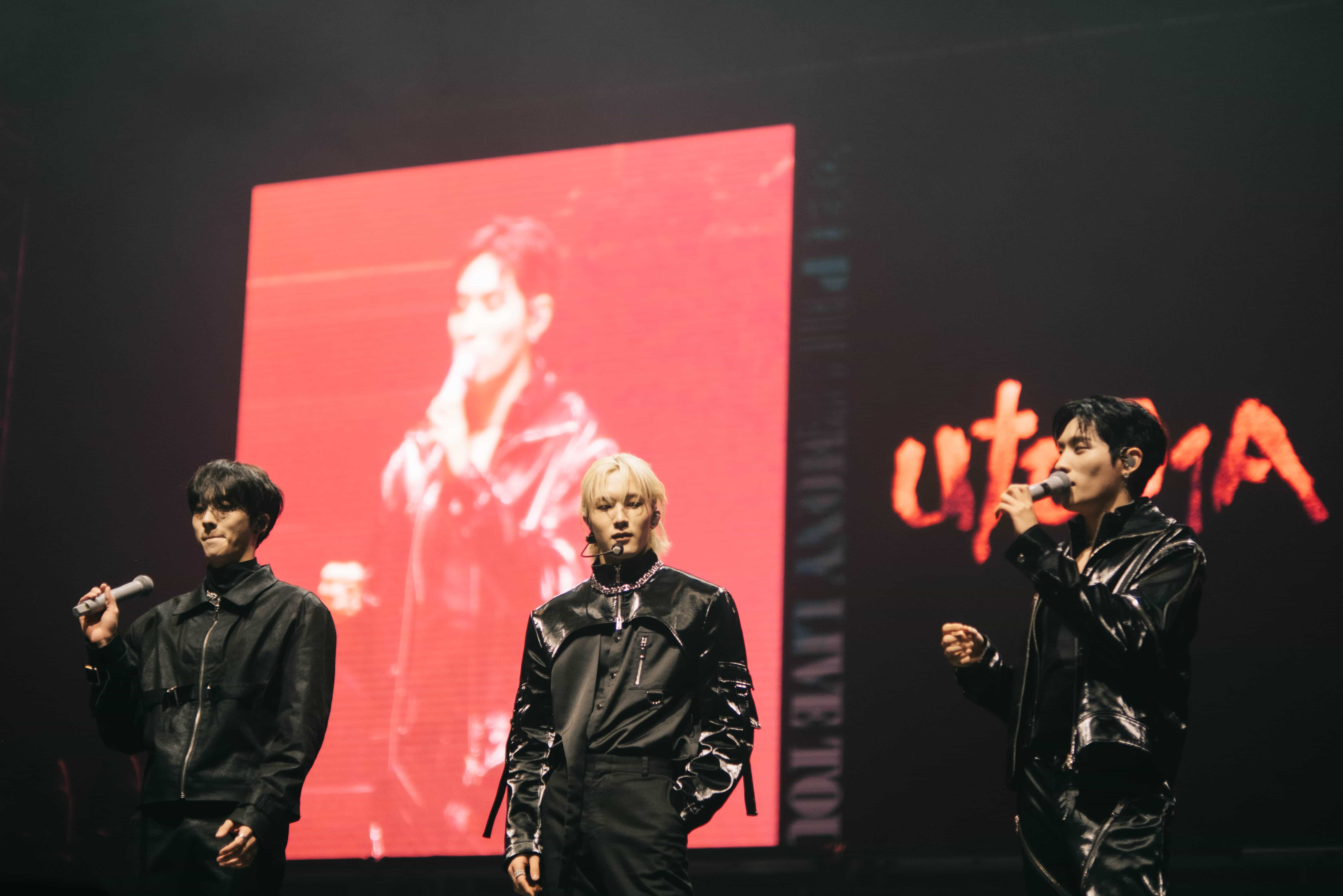 Three P1Harmony members in black leather talking to the crowd in front of a red UTOP1A screen