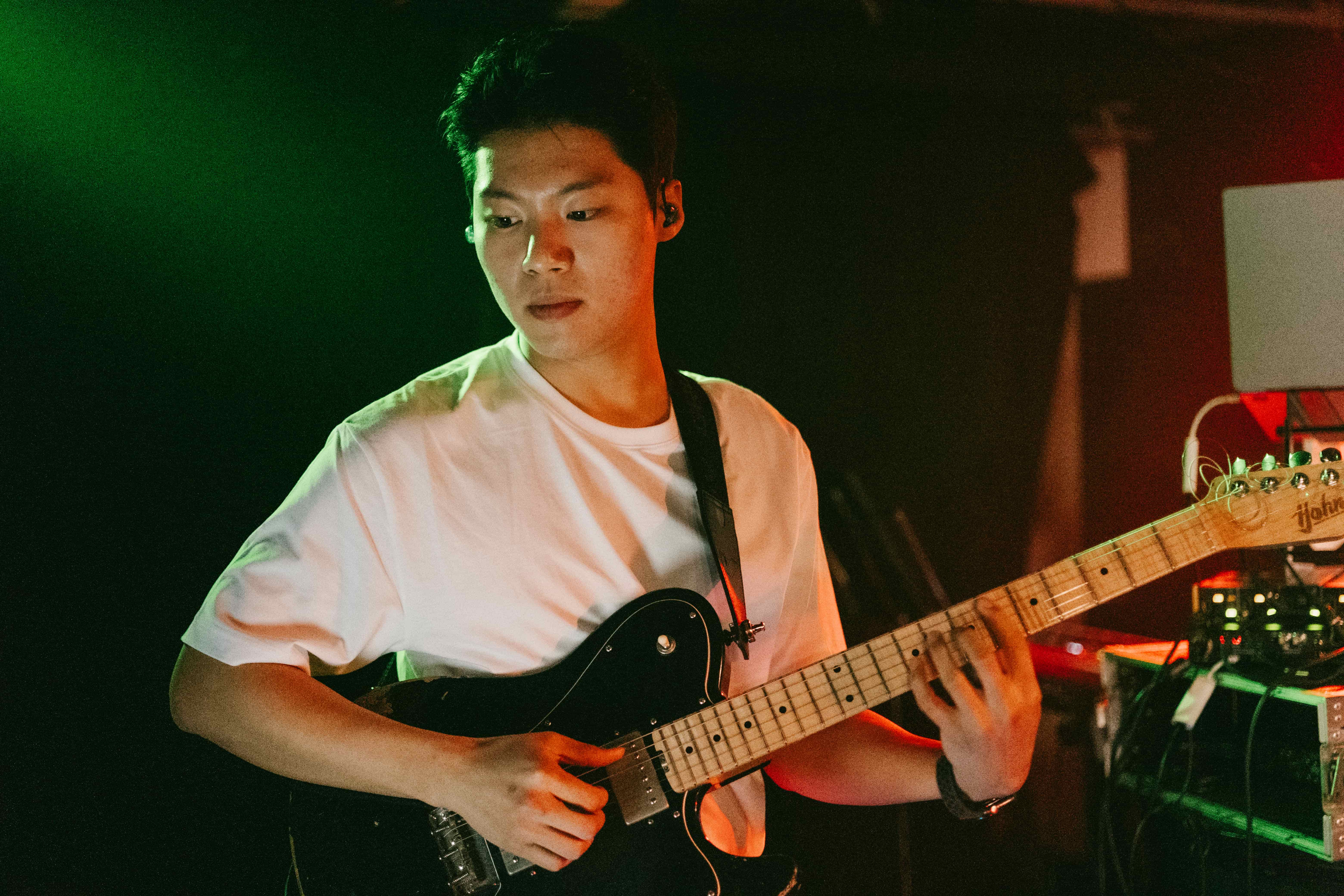 Guitarist in a white t-shirt playing a black electric guitar under green and red stage light