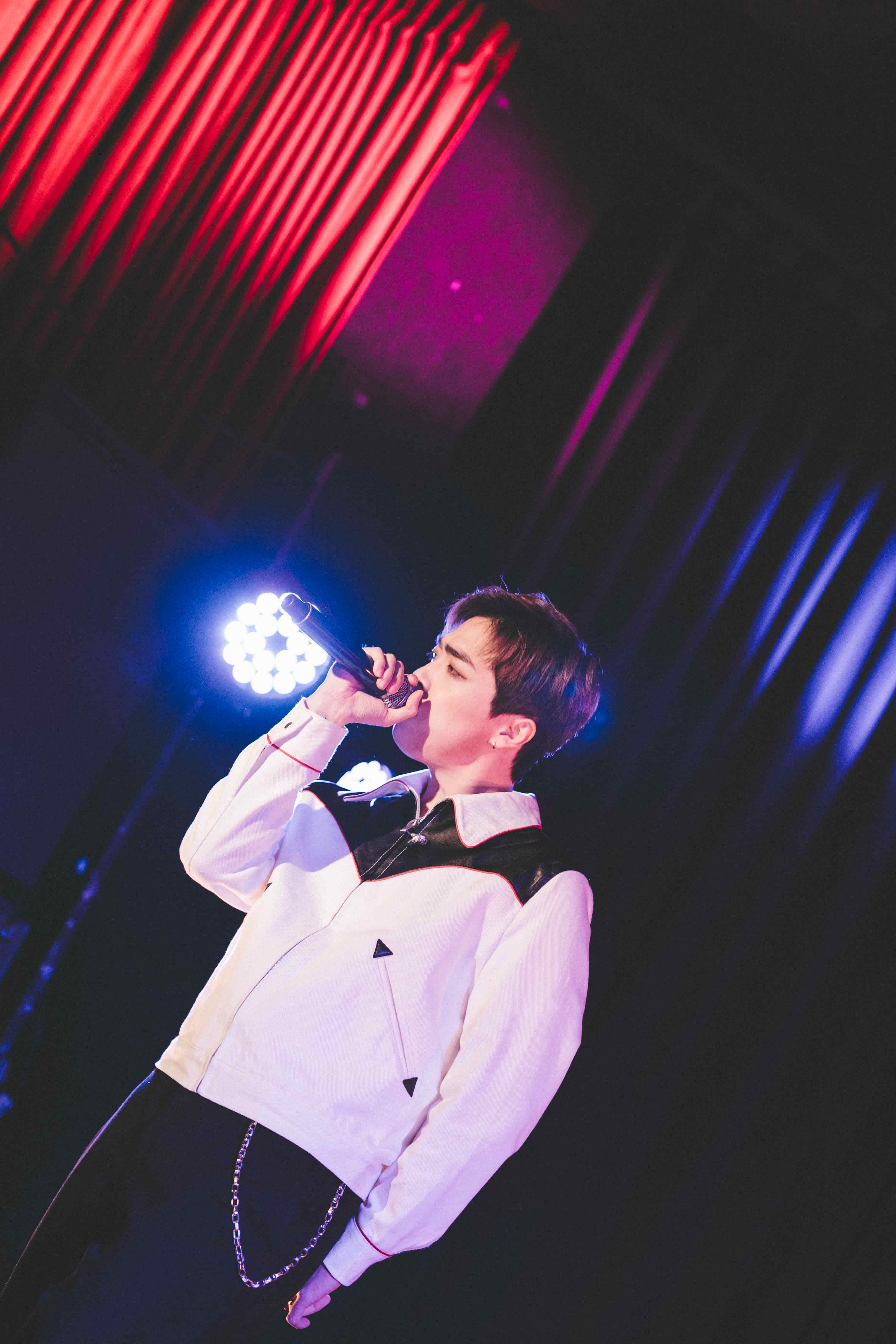 Junny singing under a bright blue spotlight with a red curtain glowing above the stage