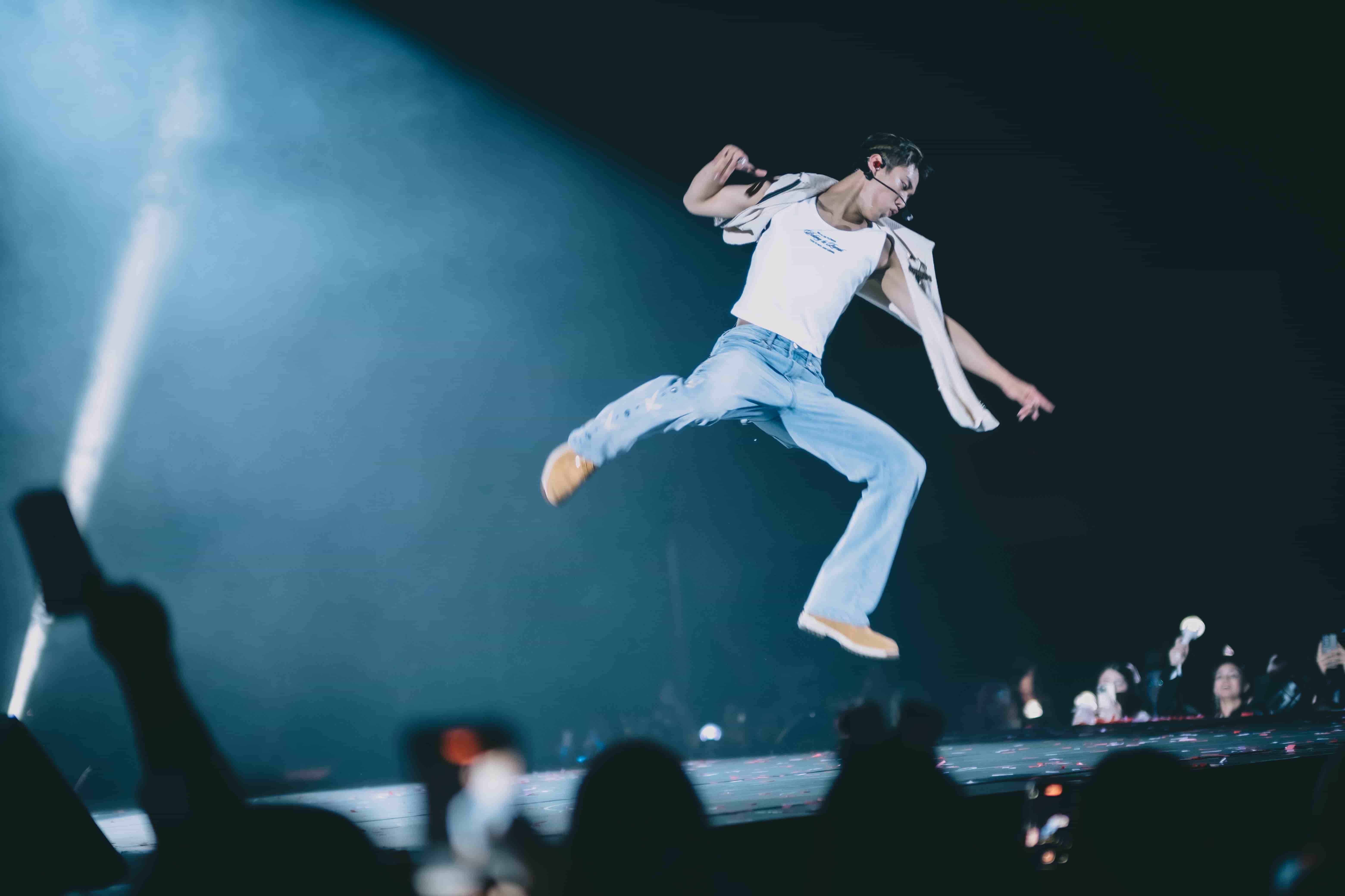 P1Harmony member jumping mid-air in light jeans and a white top above fans holding up phones