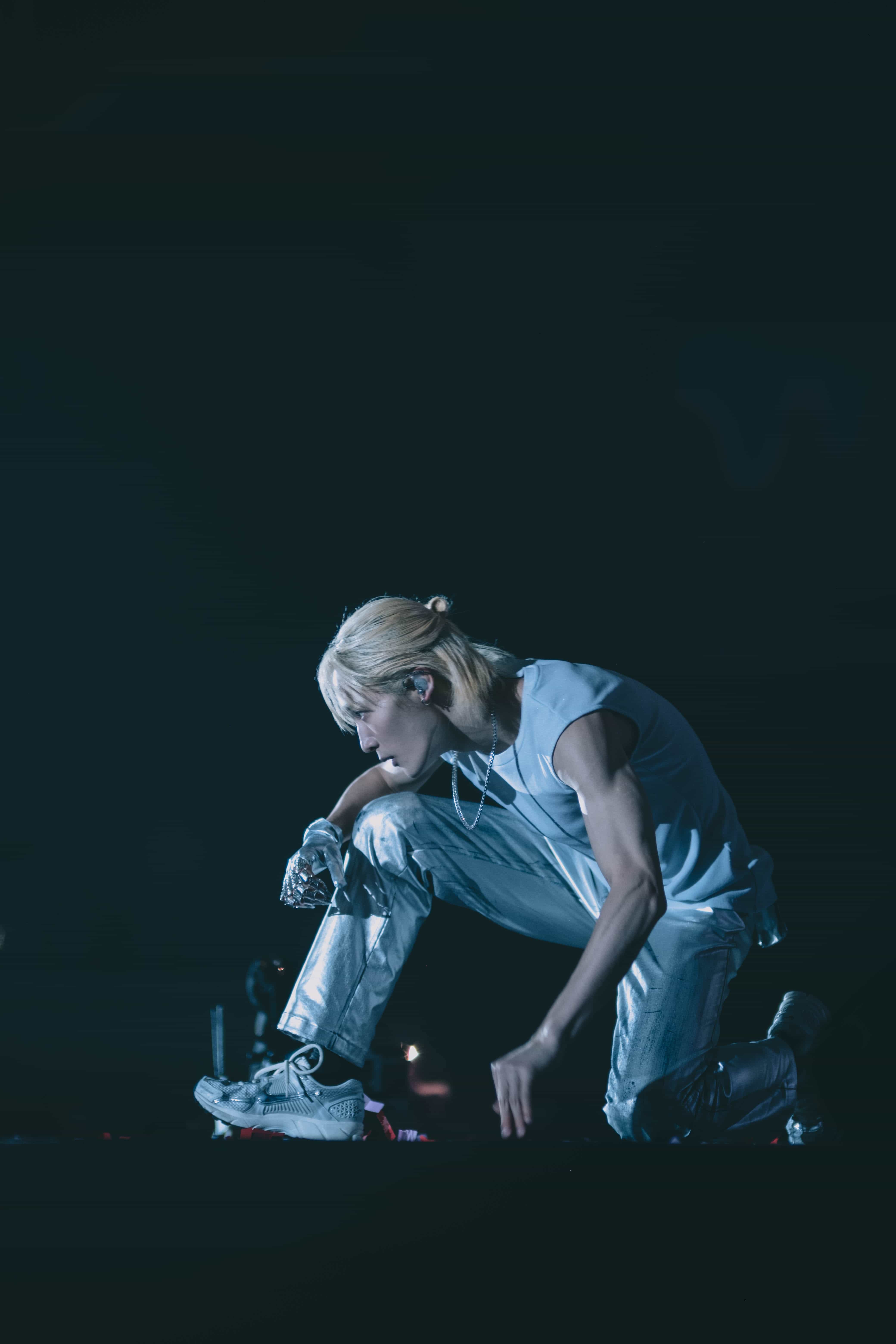 P1Harmony member with bleached hair kneeling low on stage in a silver outfit under blue light