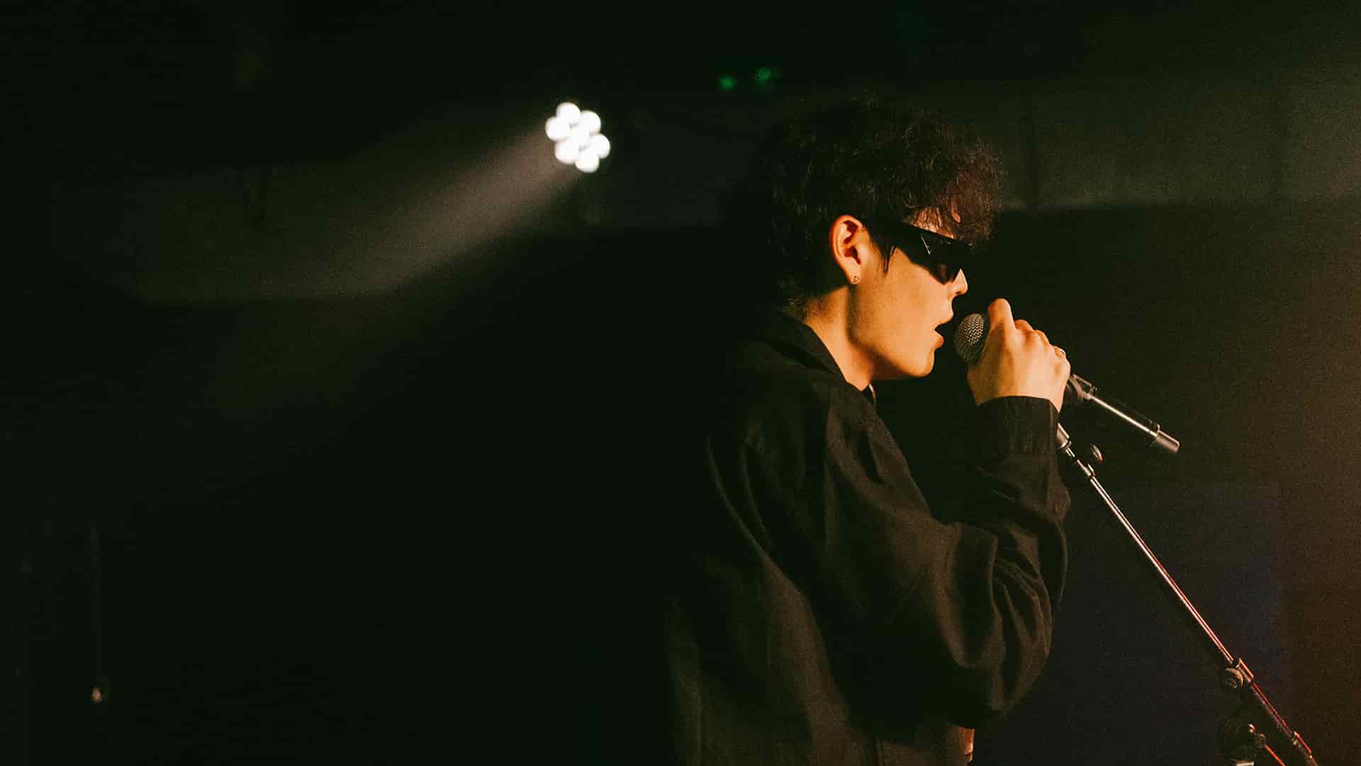 Jimmy Brown in sunglasses singing into a microphone on a dark stage under a single spotlight