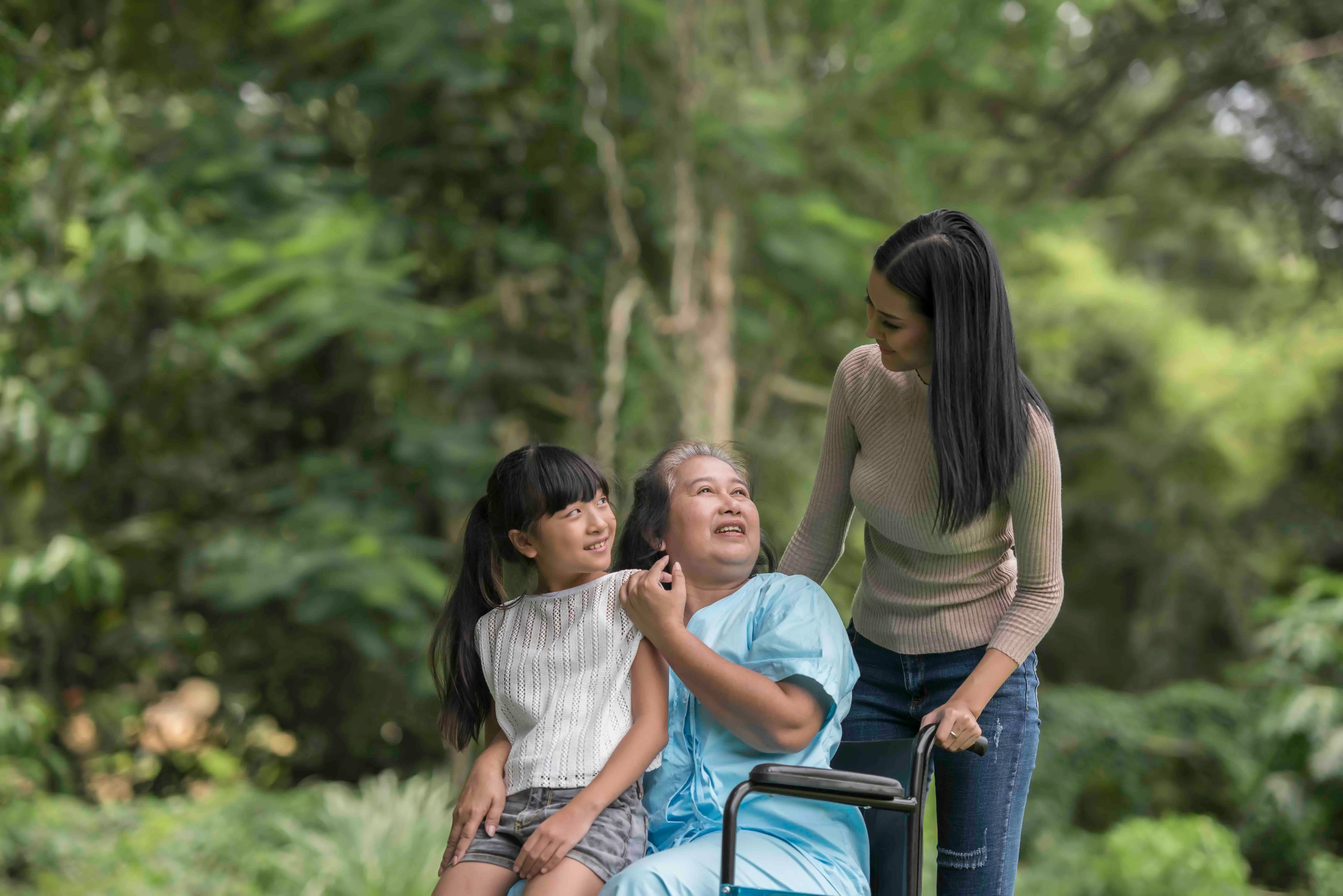 An elderly woman in a wheelchair sits outdoors in a lush, green park, smiling and holding hands with a young girl sitting on her lap.