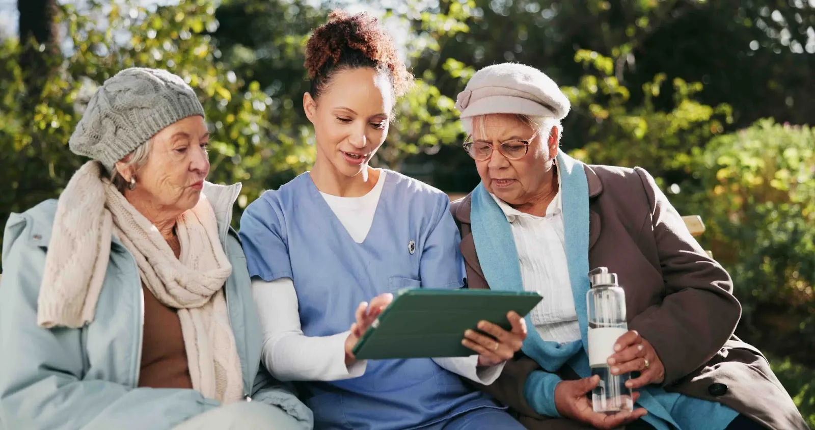 A caregiver supports two elderly women by using Caily, a caregiver app, on a tablet, demonstrating the use of digital tools in in-home senior care and assisted living settings.