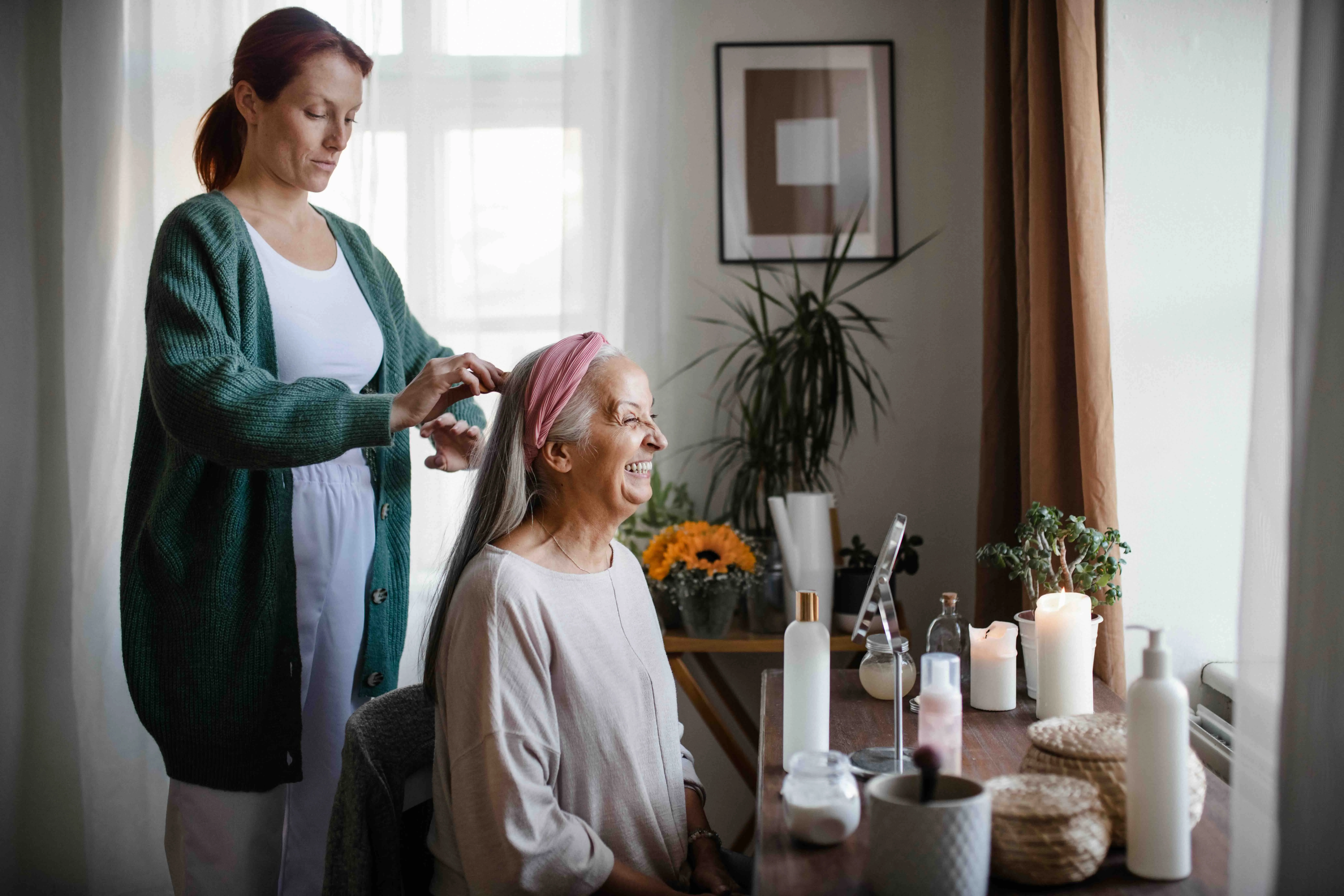 A caregiver providing home care by helping an elderly woman with her hair at home, illustrating the warmth and dignity of in-home care services and home care elder care. Candles, plants, and self-care items on the table emphasize a peaceful at home care e