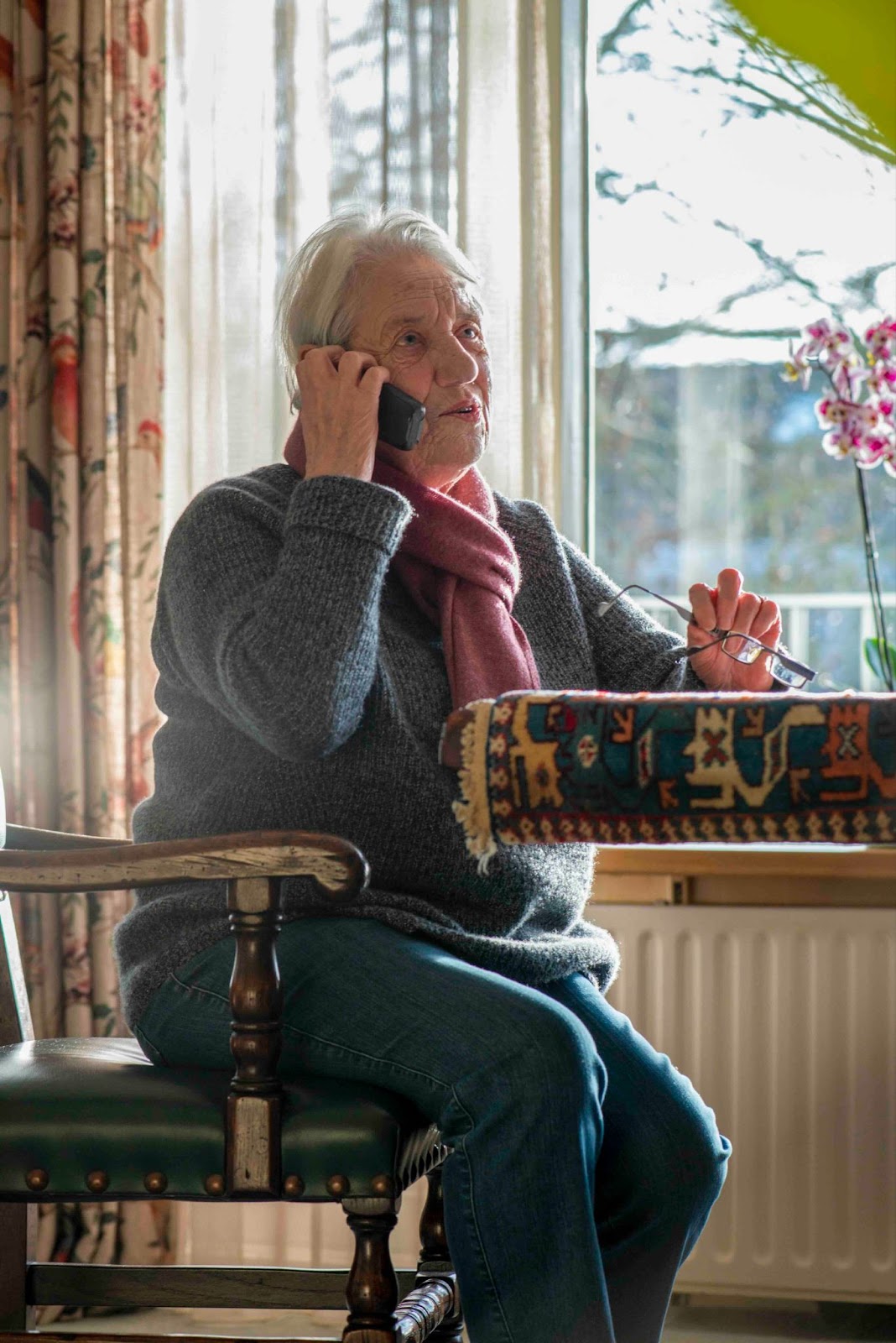Older woman sitting at home, talking on the phone—possibly calling an airline about a bereavement flight or seeking travel assistance after a family loss.