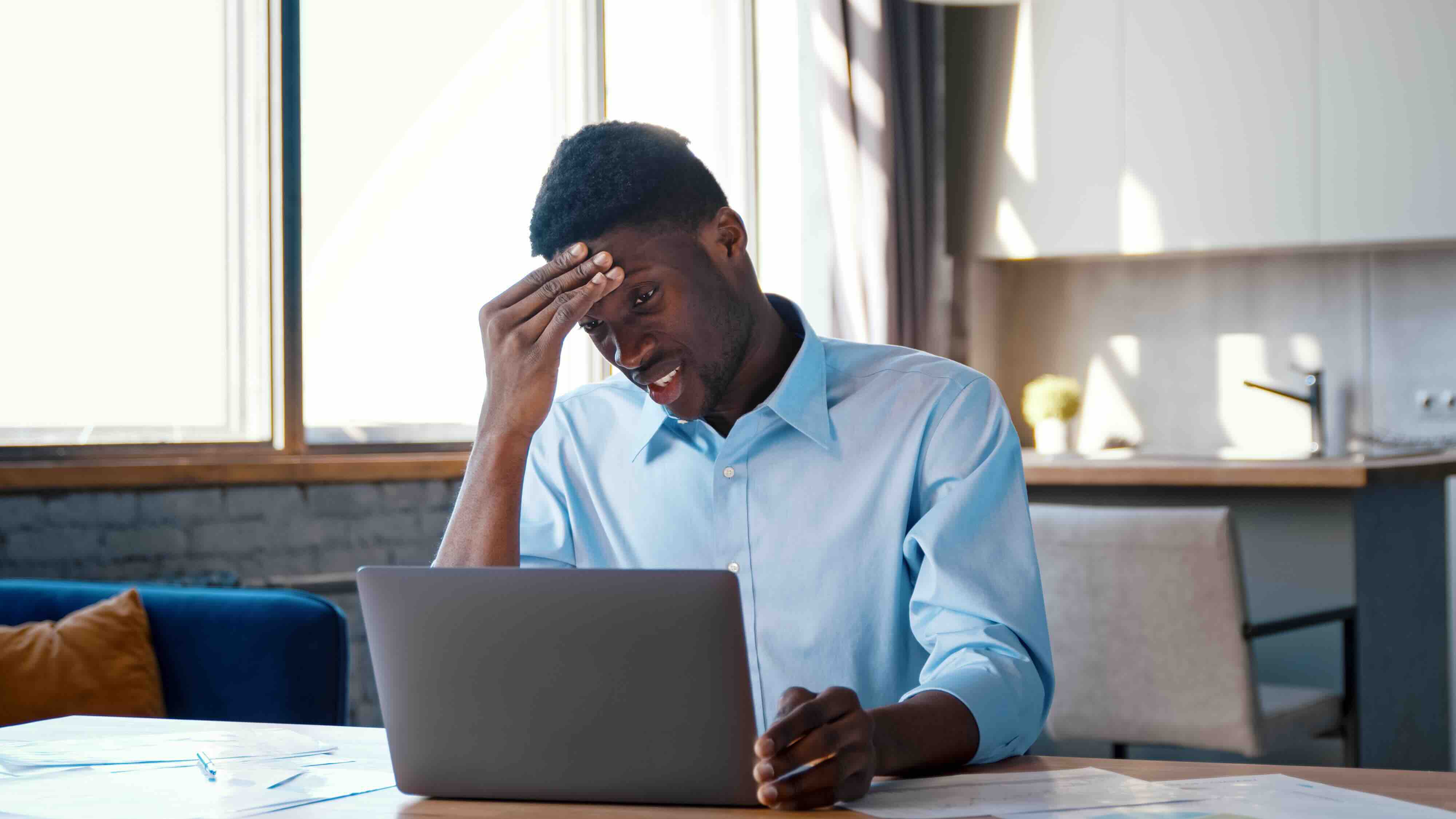 A man sits at a table looking stressed while using a laptop, possibly managing subscription cancellation after death or closing accounts after death. Papers are spread out in front of him, suggesting he's working through a checklist of things to cancel whe
