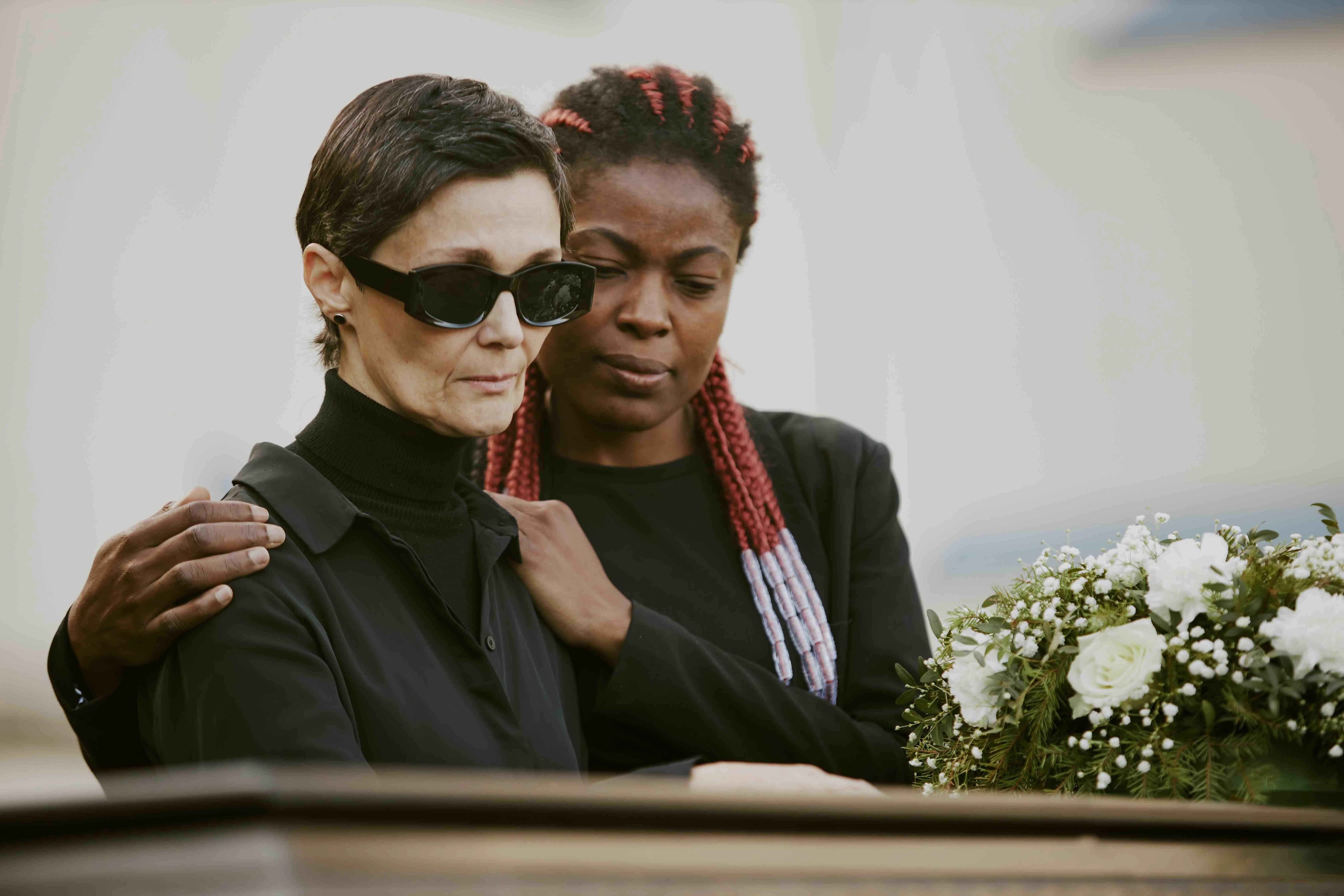 Two women dressed in black stand beside a casket adorned with white flowers, comforting each other during a funeral ceremony—capturing the emotional weight often present when delivering a eulogy or funeral speech.