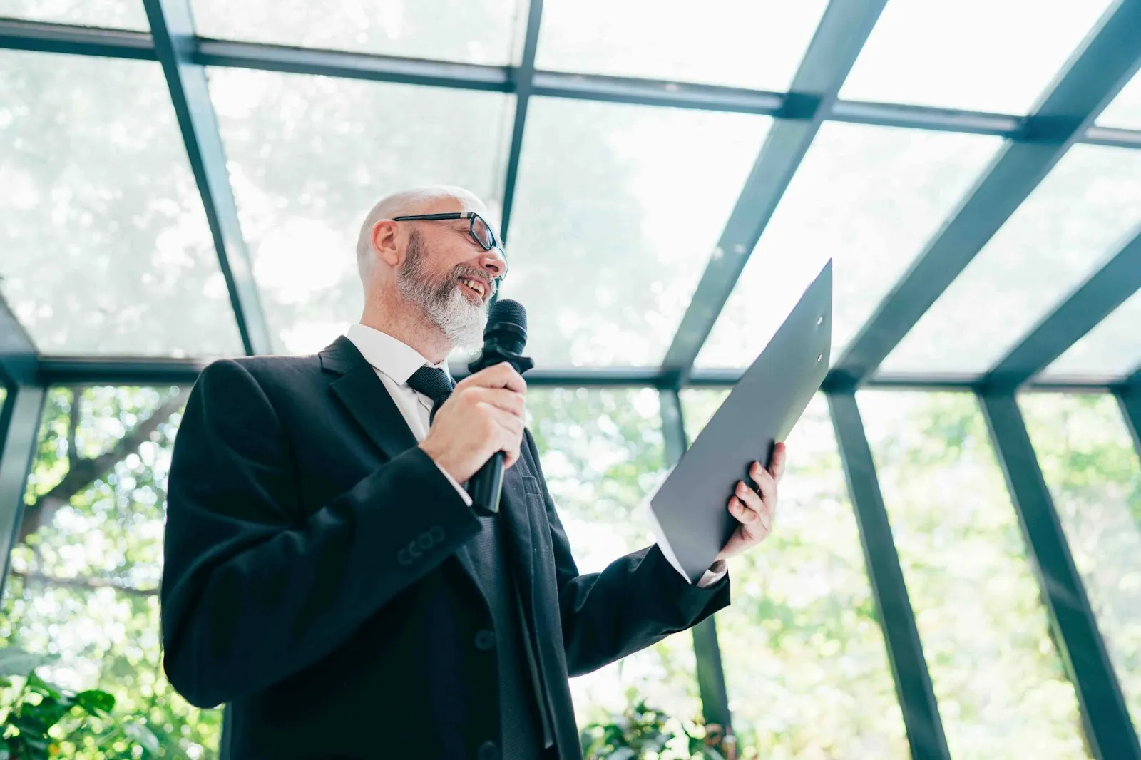 A man in a suit delivers a funeral speech, holding a microphone and reading from a folder during a memorial service, representing a moment of connection through a heartfelt eulogy.