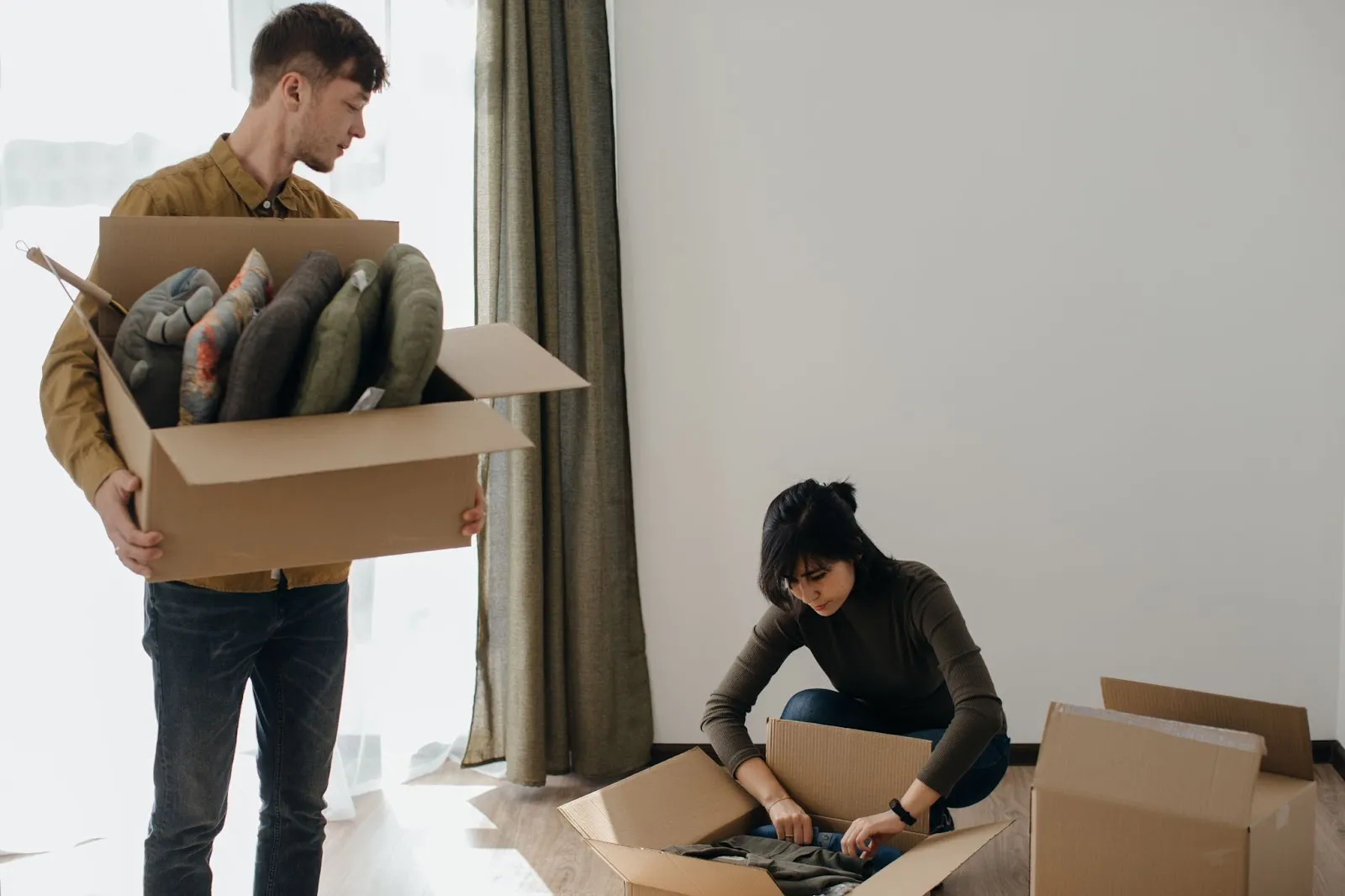 Man carrying a box of cushions while woman unpacks clothing during cleaning out parents’ house after death as part of an estate cleanout process.
