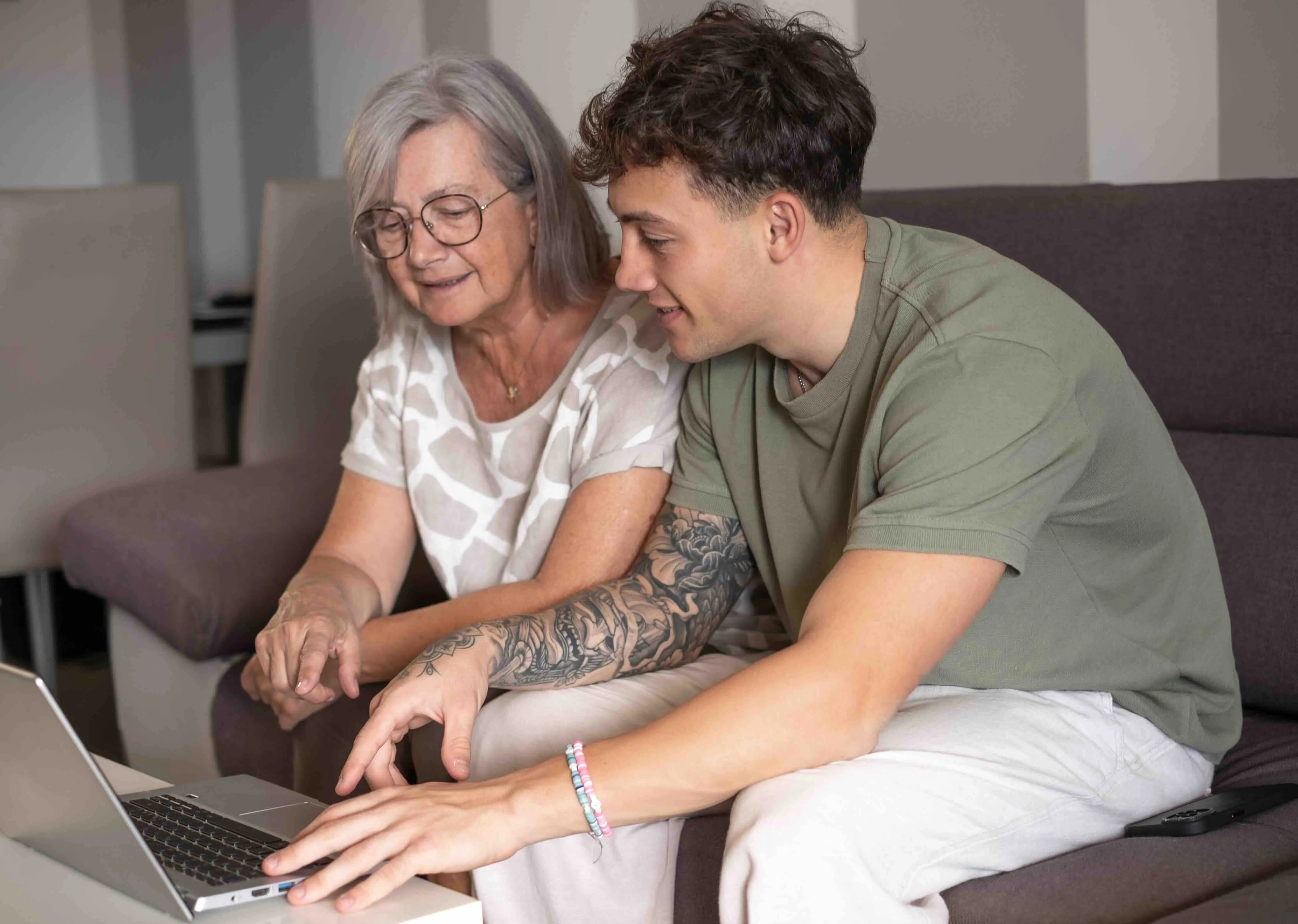 A young man helps an older woman use a laptop at home, symbolizing family caregiving and intergenerational support. This reflects the value of empathy and support for caregivers in creating a positive work environment and boosting employee retention.