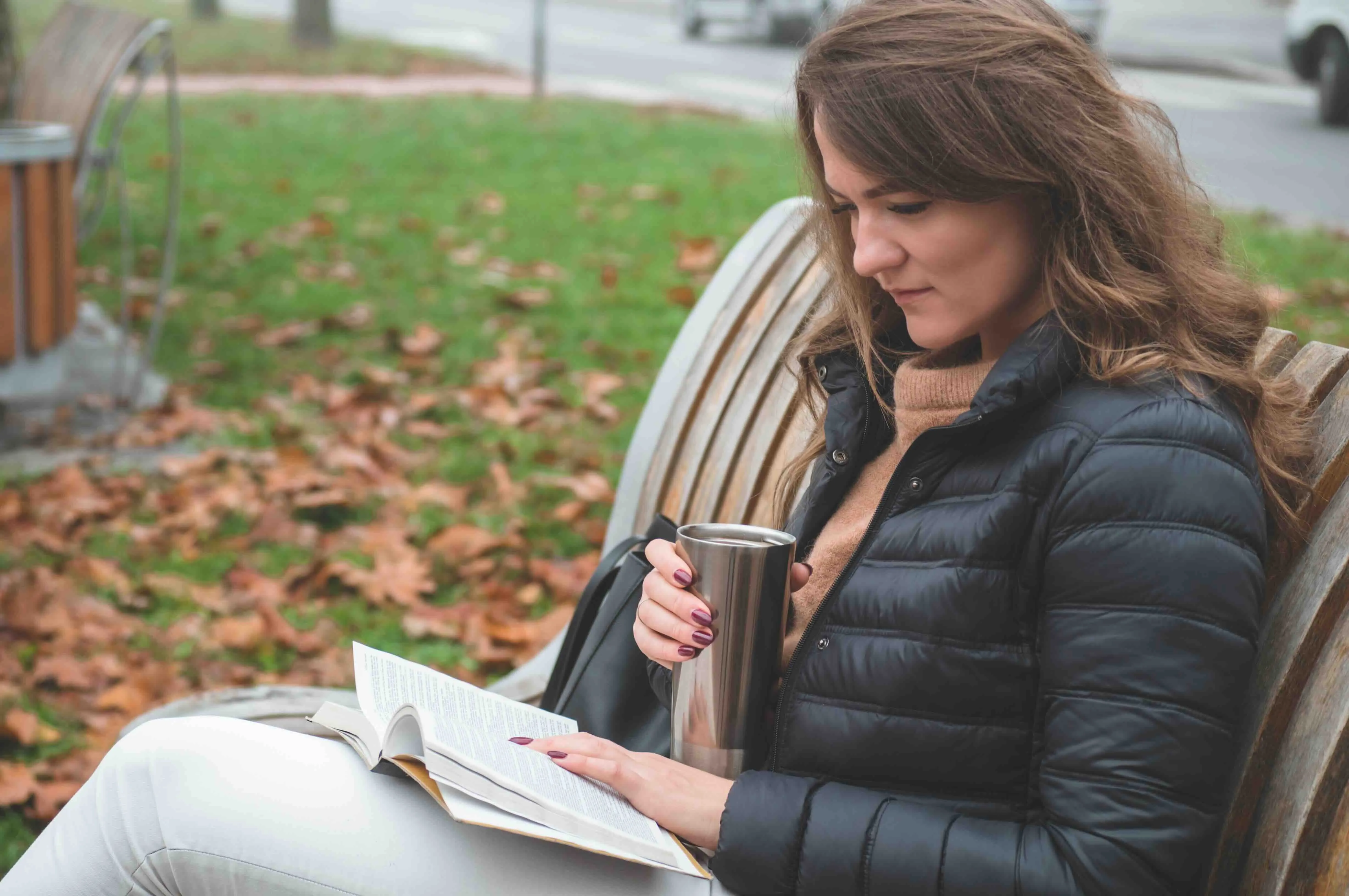 Woman reading a book about grief while sitting on a park bench in autumn, holding a coffee cup — books for grief, talking to kids about death, and explaining death to children.
