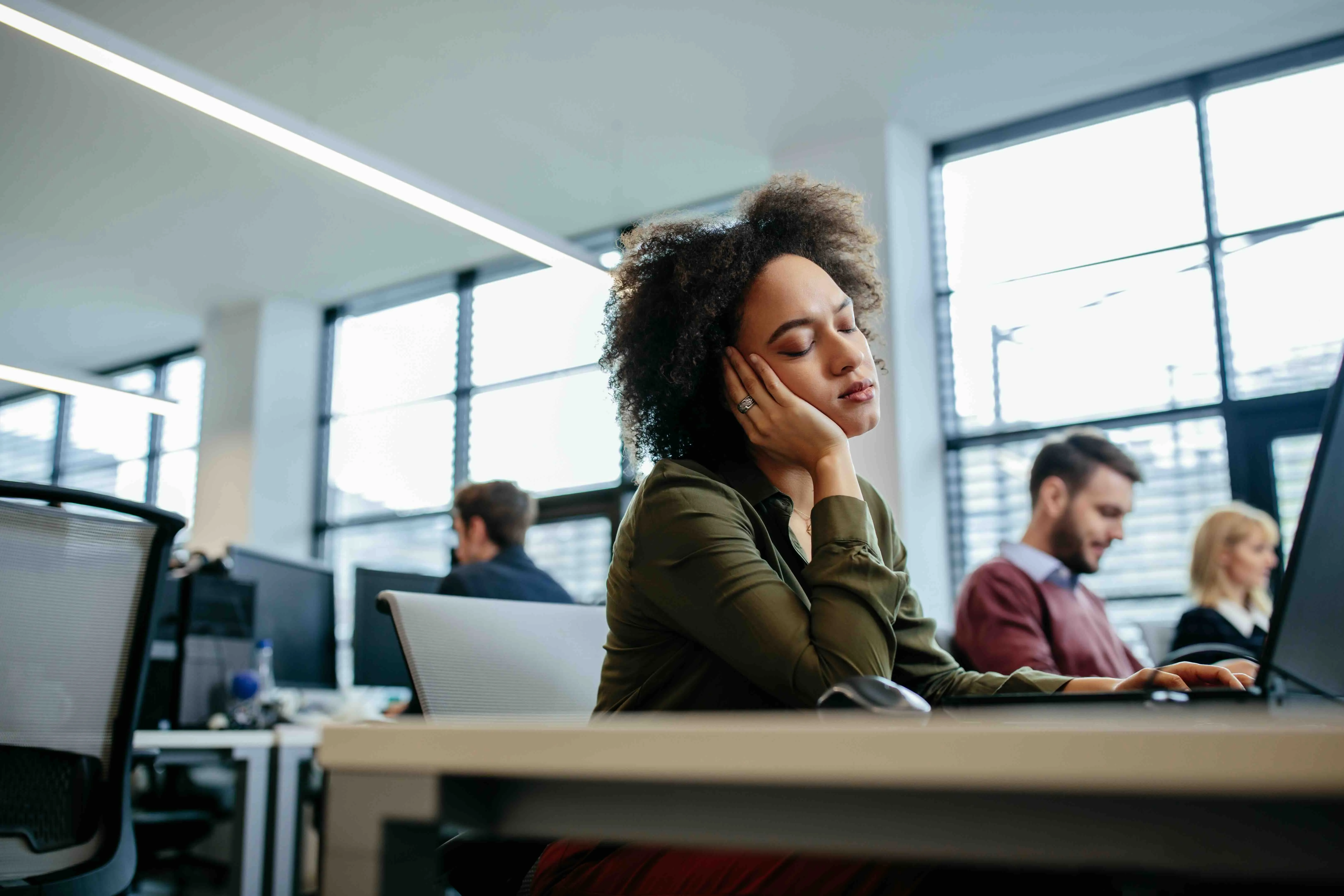 A tired employee sitting at an office desk with her head resting on her hand, eyes closed, appearing exhausted while working on a laptop — symbolizing caregiver burnout in the workplace.