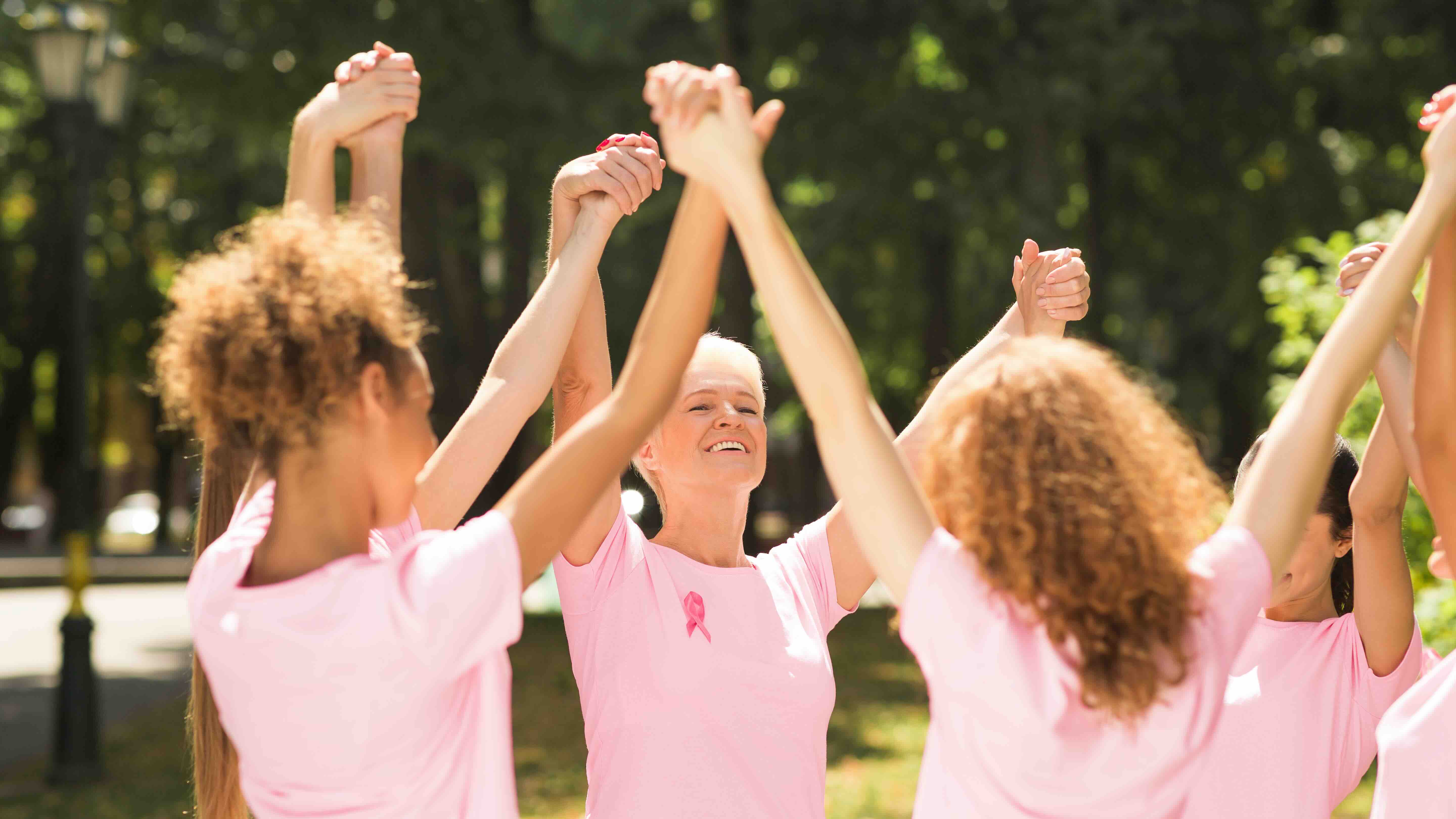 Group of women wearing pink shirts with ribbons holding hands in support during Breast Cancer Awareness Month.