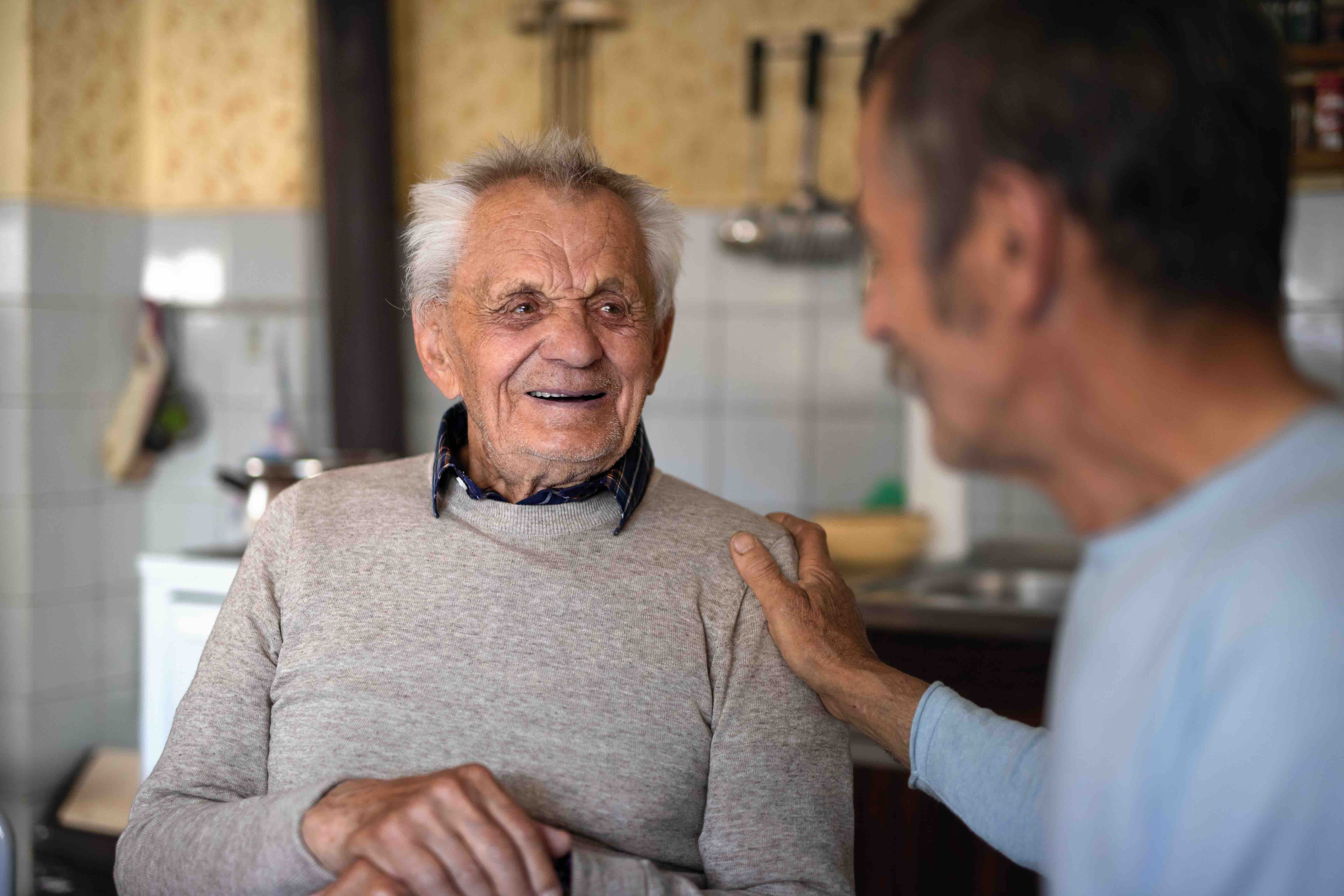 An elderly father and his adult son share a warm moment together in a cozy kitchen, smiling and talking while preserving their family history through meaningful conversation and connection.