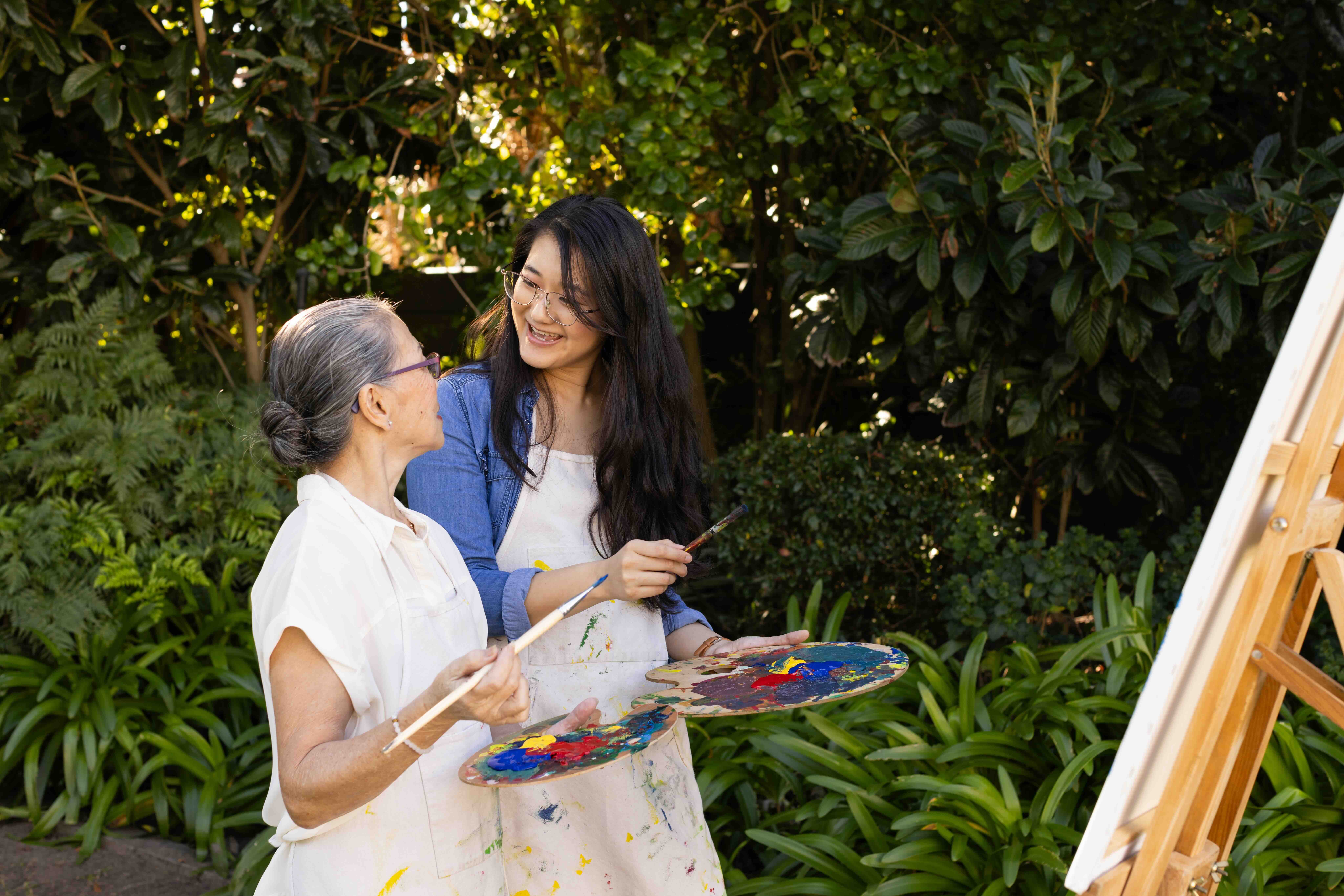 A caregiver and an older loved one painting together outdoors, smiling and sharing a creative moment surrounded by greenery. This photo represents connection, joy, and balance in caregiving — emotional labor, caregiver burnout, and carers’ mental health.'