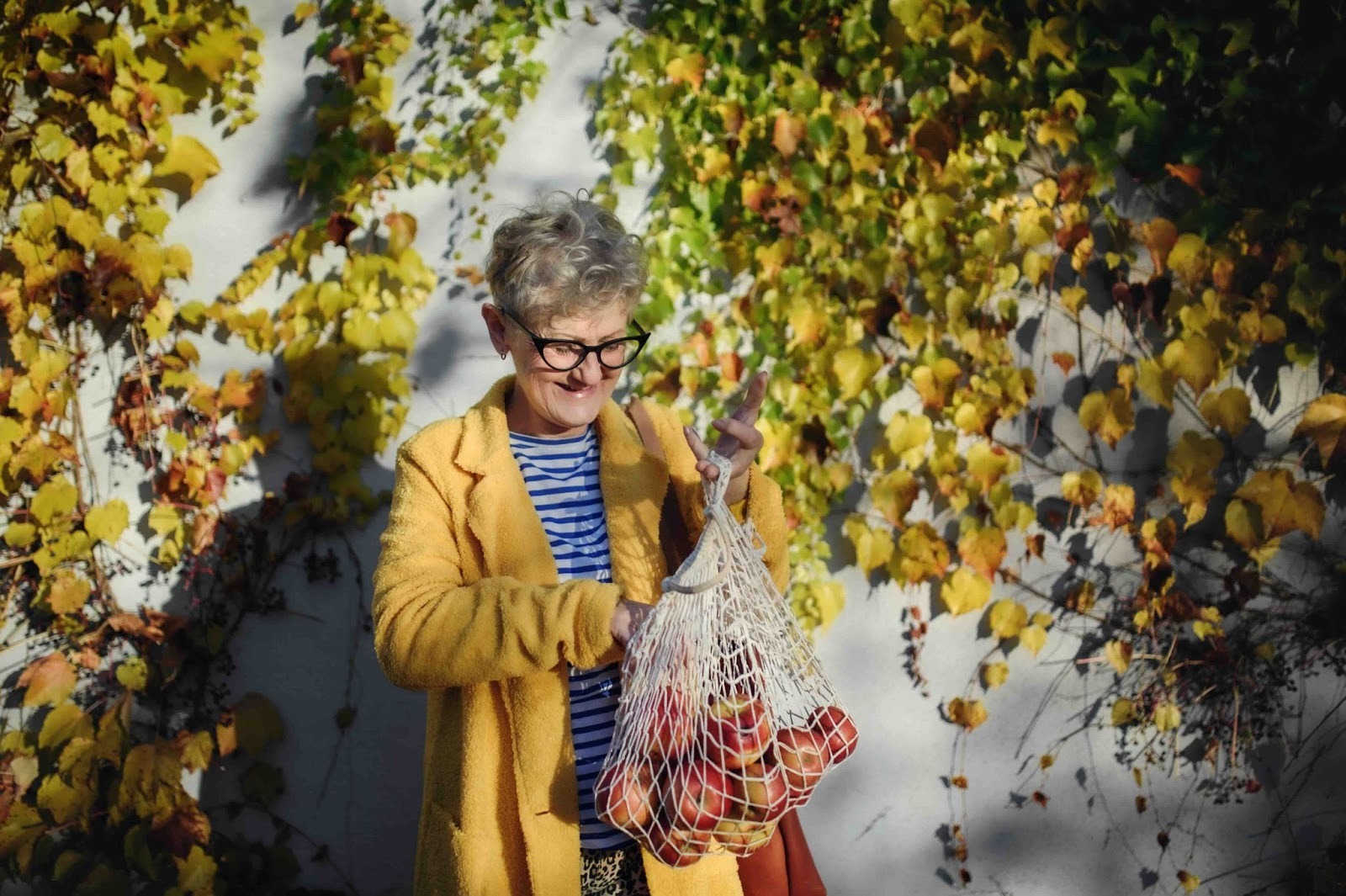 Older woman smiling outdoors in autumn, holding a net bag of apples against colorful fall leaves — representing a calm, seasonal moment in dementia care and a dementia friendly approach to Halloween 2025 activities.