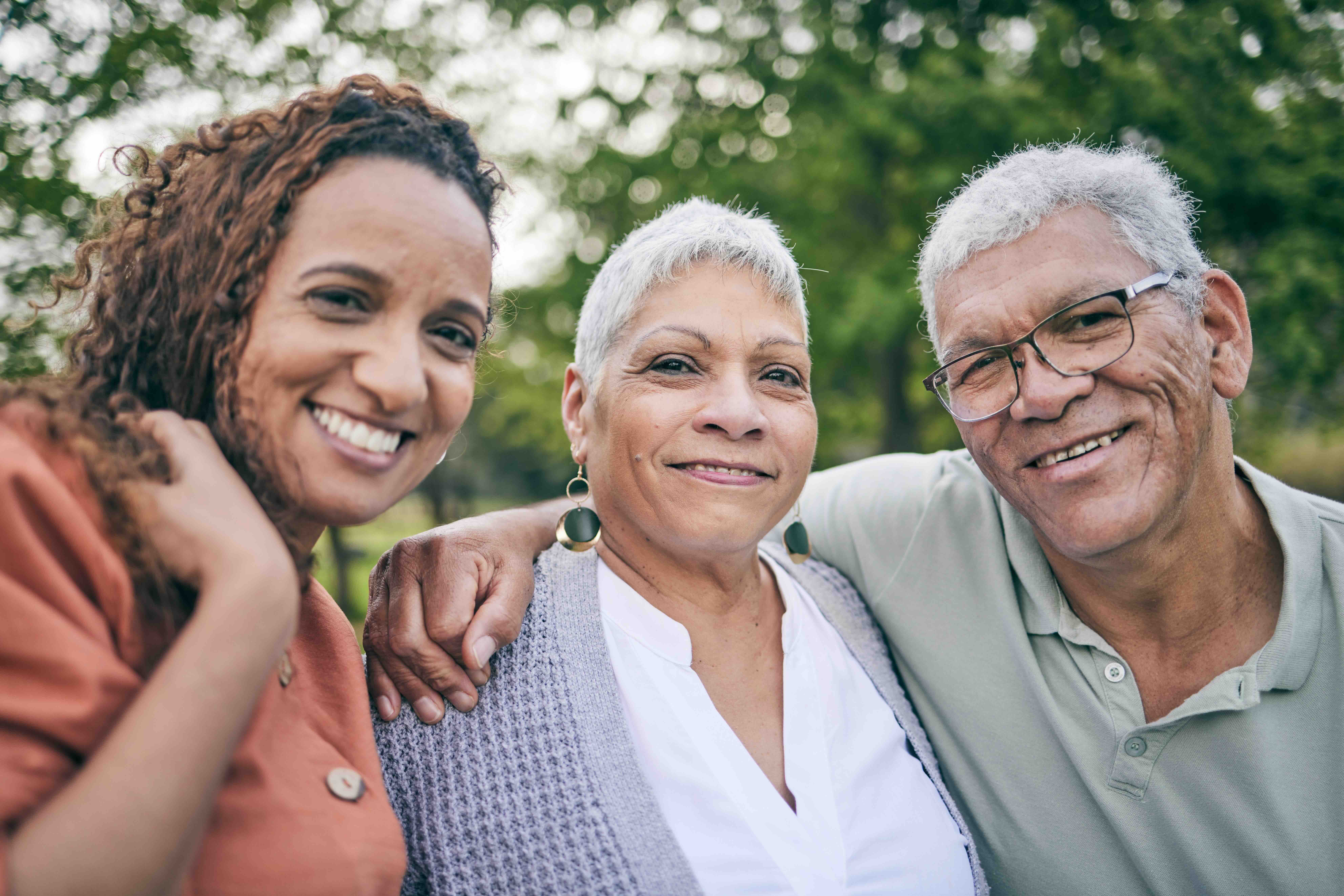  A smiling woman with her arm around two older adults, standing outdoors together and symbolizing the warmth, connection, and support of family caregiving during National Family Caregiver Awareness Month 2025