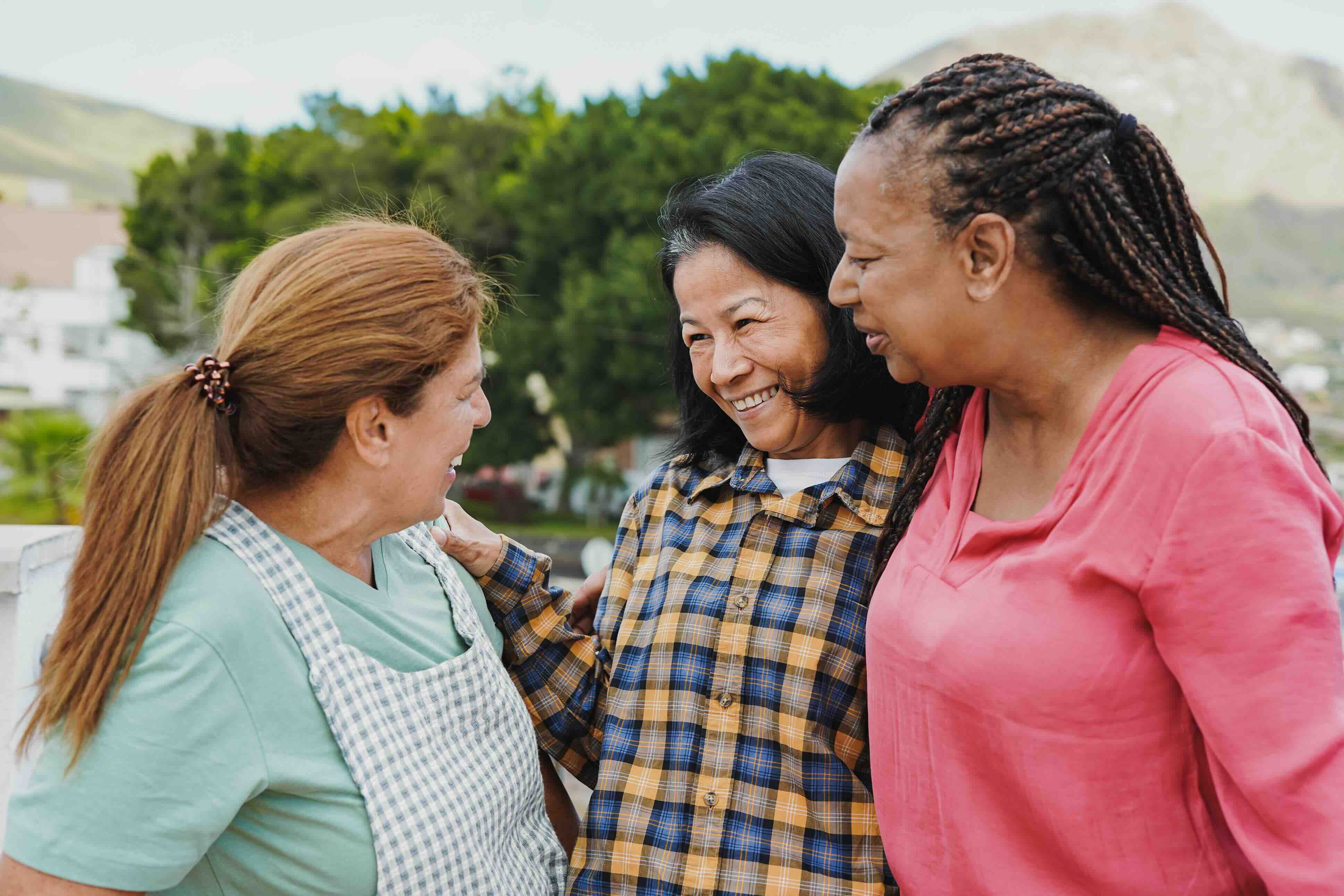 Three women from different backgrounds stand together outdoors, smiling and supporting one another, representing connection and community found in caregiver support groups and support groups for caregivers