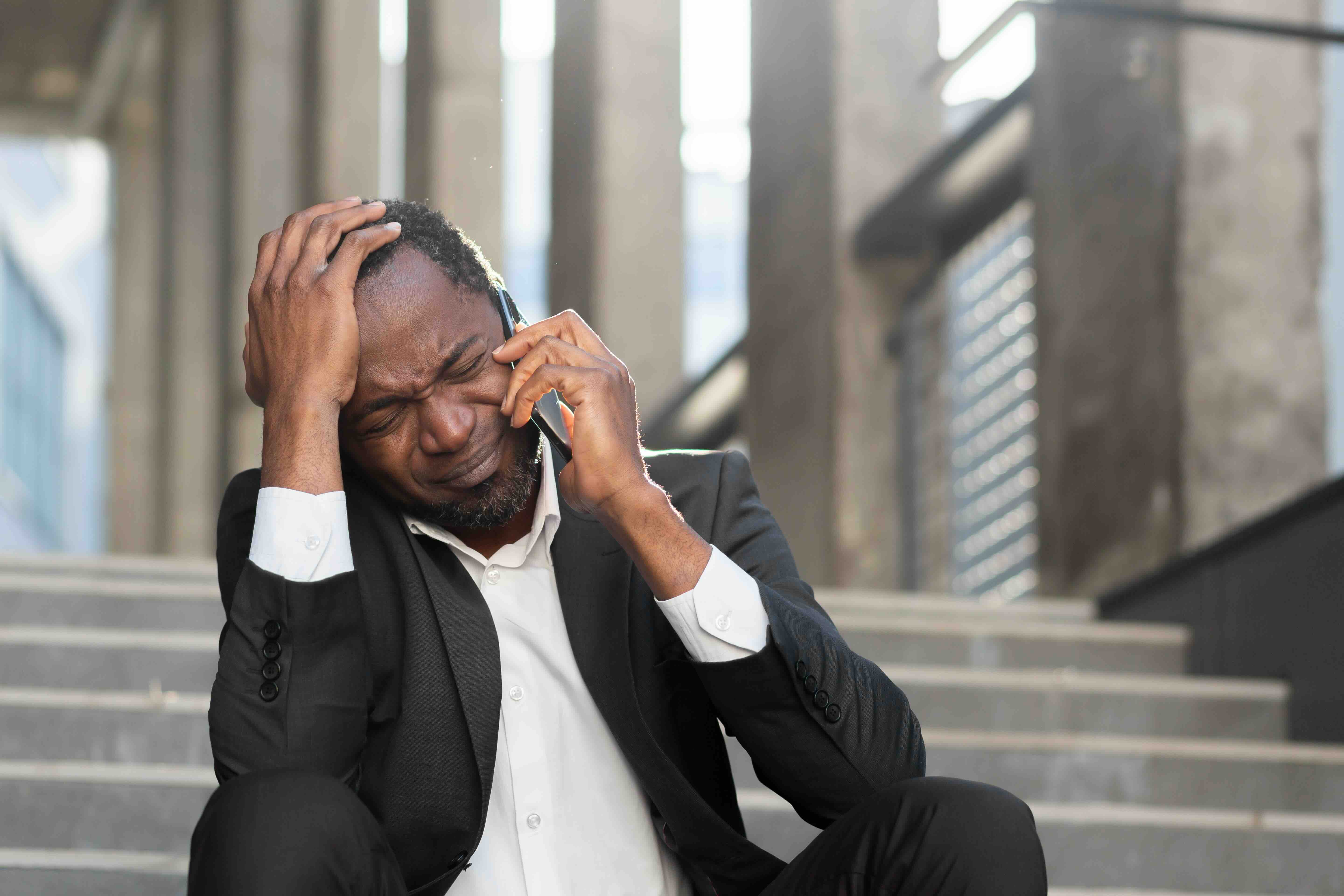 A distressed man sitting on outdoor steps, holding his head while talking on the phone during an urgent situation, illustrating the emotional intensity of emergencies and the importance of knowing the emergency action steps, recognizing when medical attention is needed, and understanding the steps to follow in an emergency are essential for staying focused under pressure.