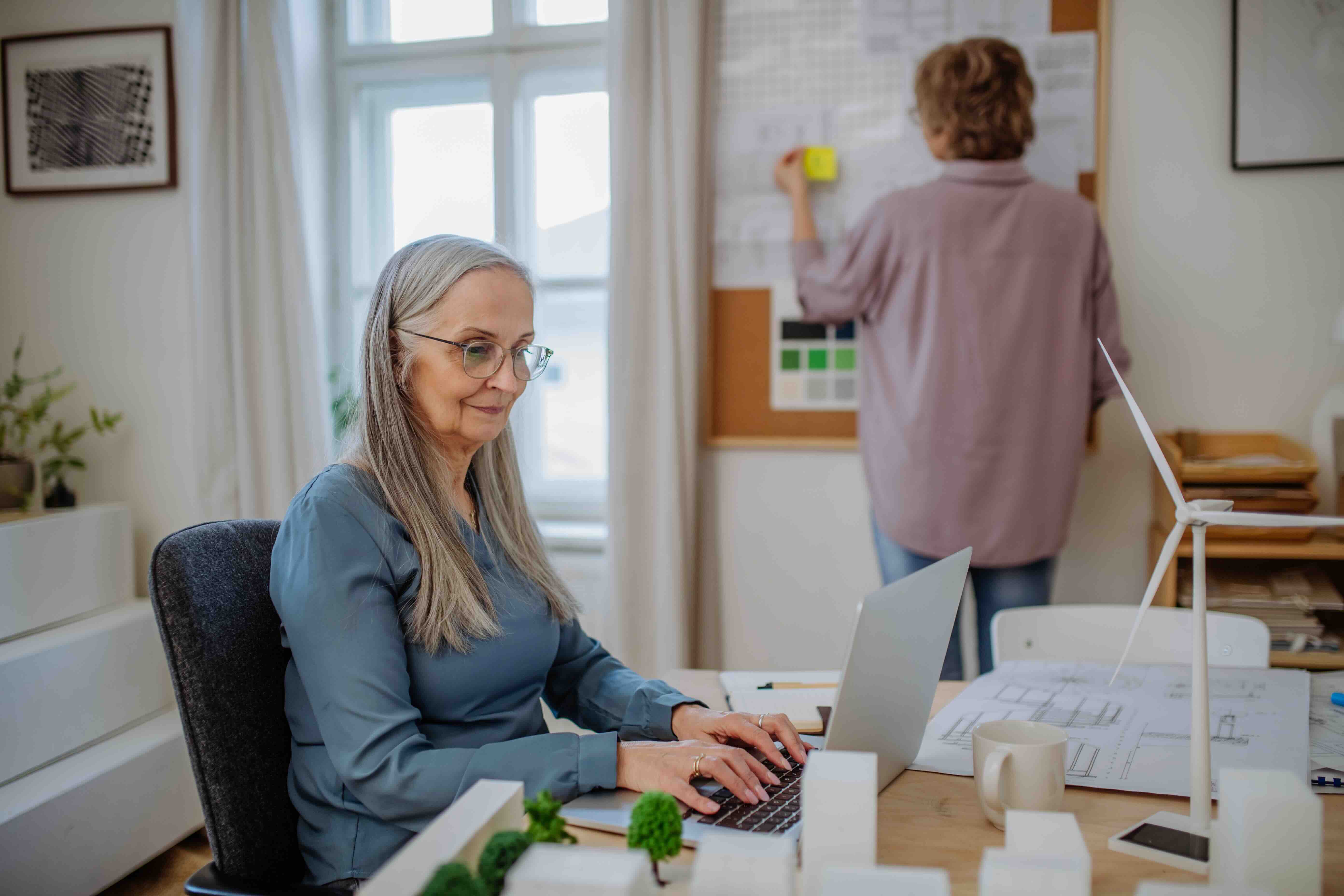 Older woman working at her laptop in a bright home office while another person reviews plans on a bulletin board, representing caregivers balancing professional work and family responsibilities.