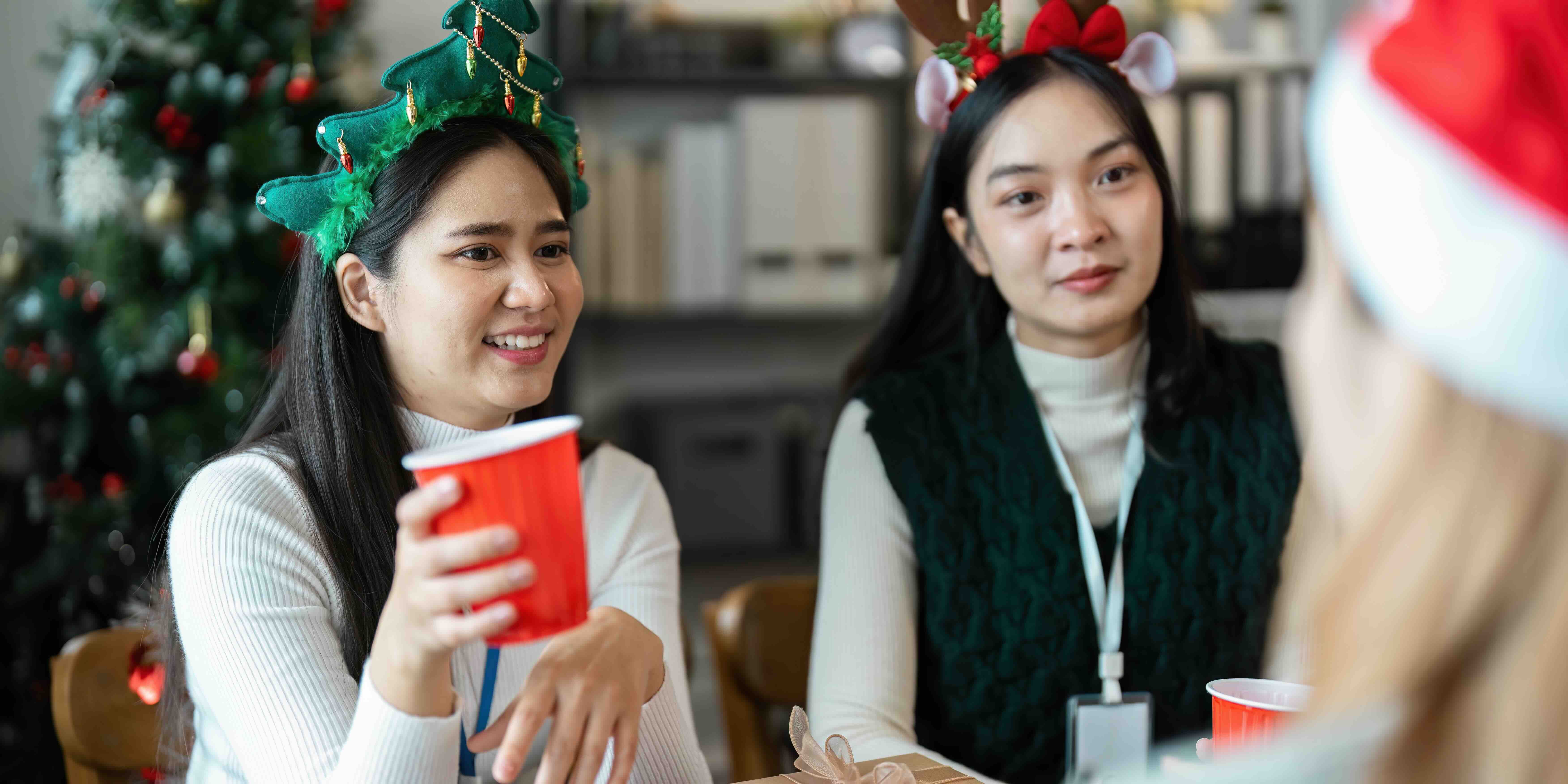 Coworkers wearing holiday accessories sit together at an office gathering, talking and sharing a light moment. The image highlights employee support during the holidays and the importance of creating a welcoming environment for every caregiver employee.