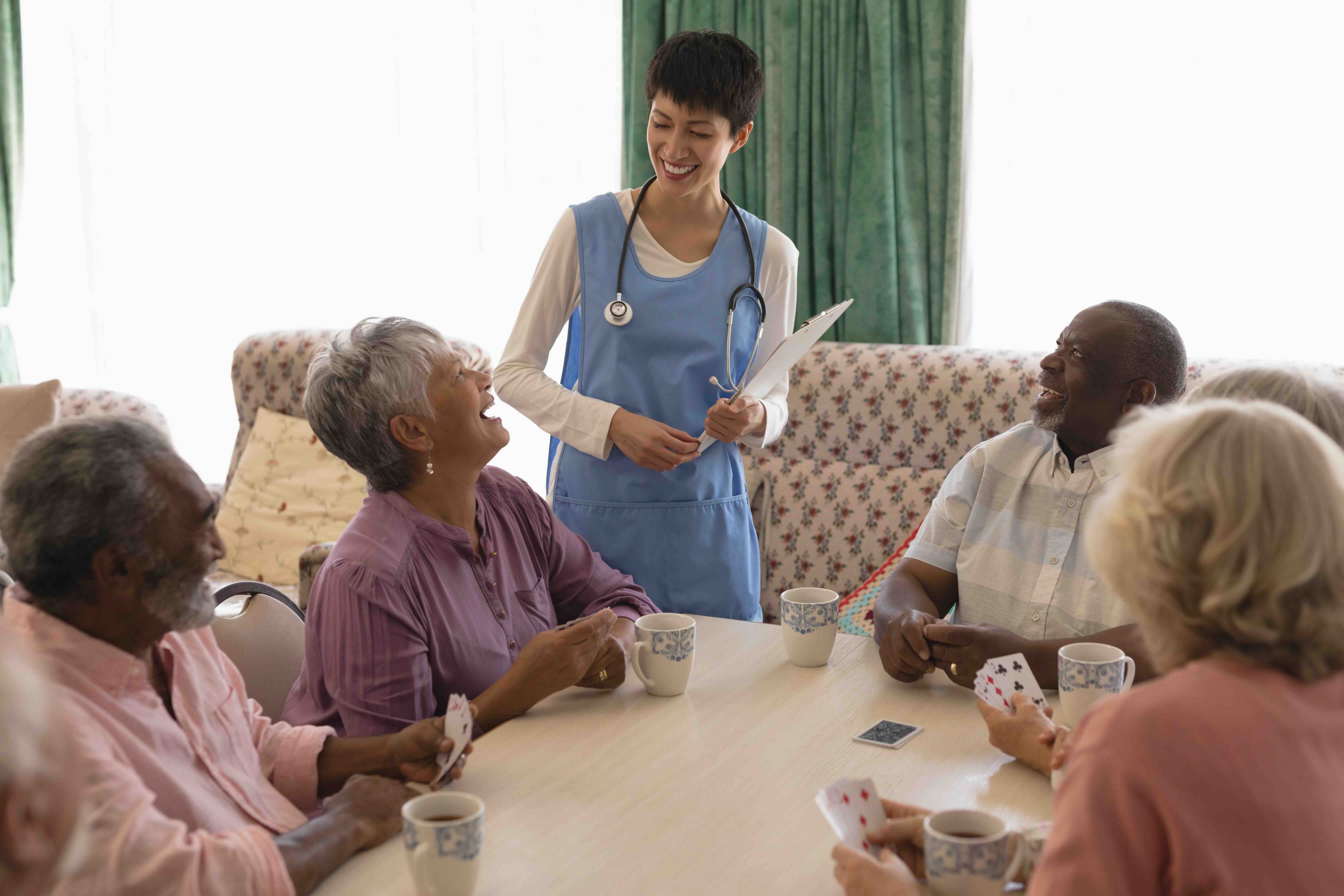 A nurse smiles while talking with a group of older adults playing cards in a communal area, illustrating the supportive environment that influences nursing home costs and the overall cost of nursing home care.