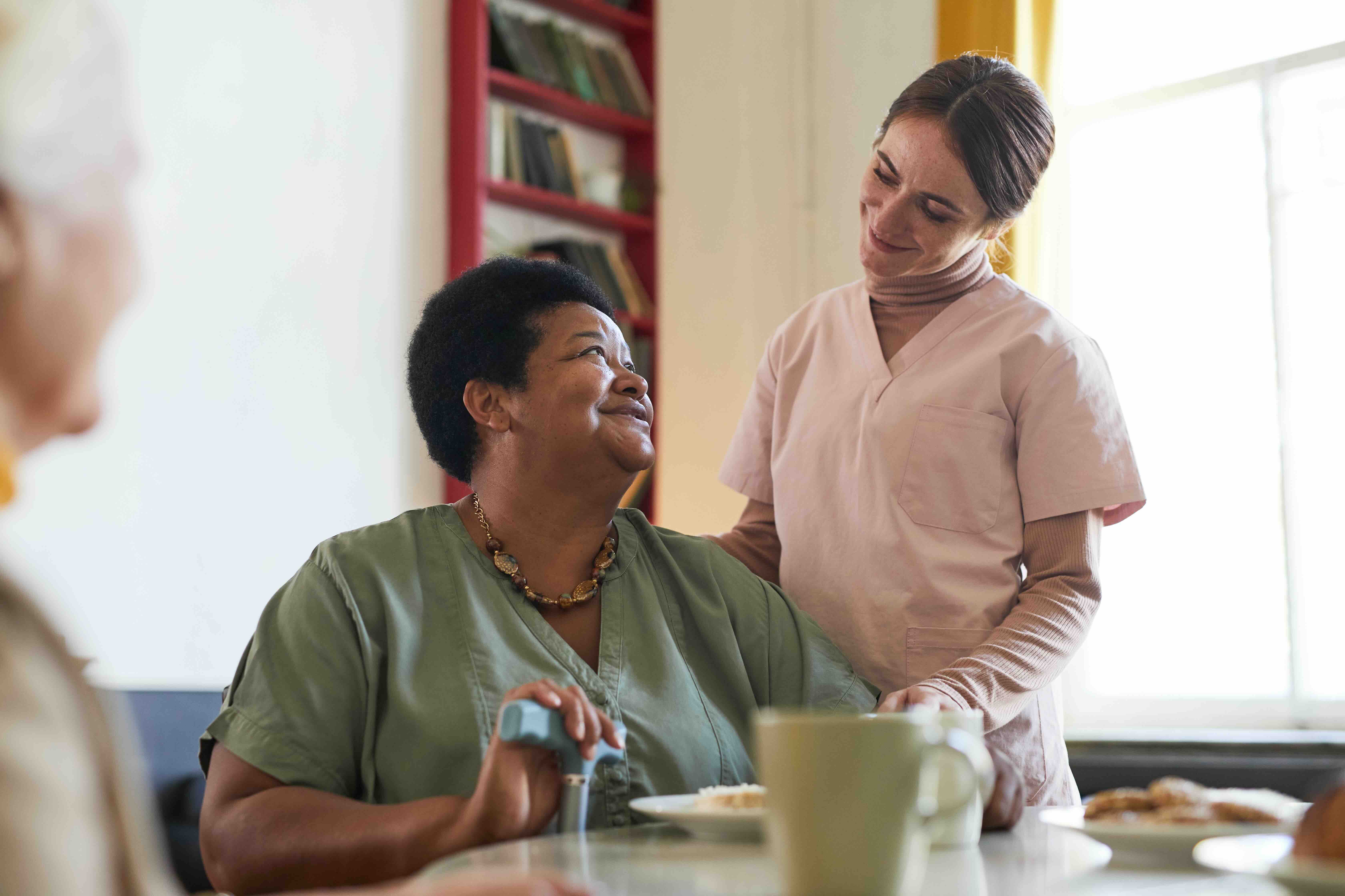A care partner smiling and checking in with a resident during mealtime at a senior living community, illustrating the warm, person-centered care culture that defines modern senior living.