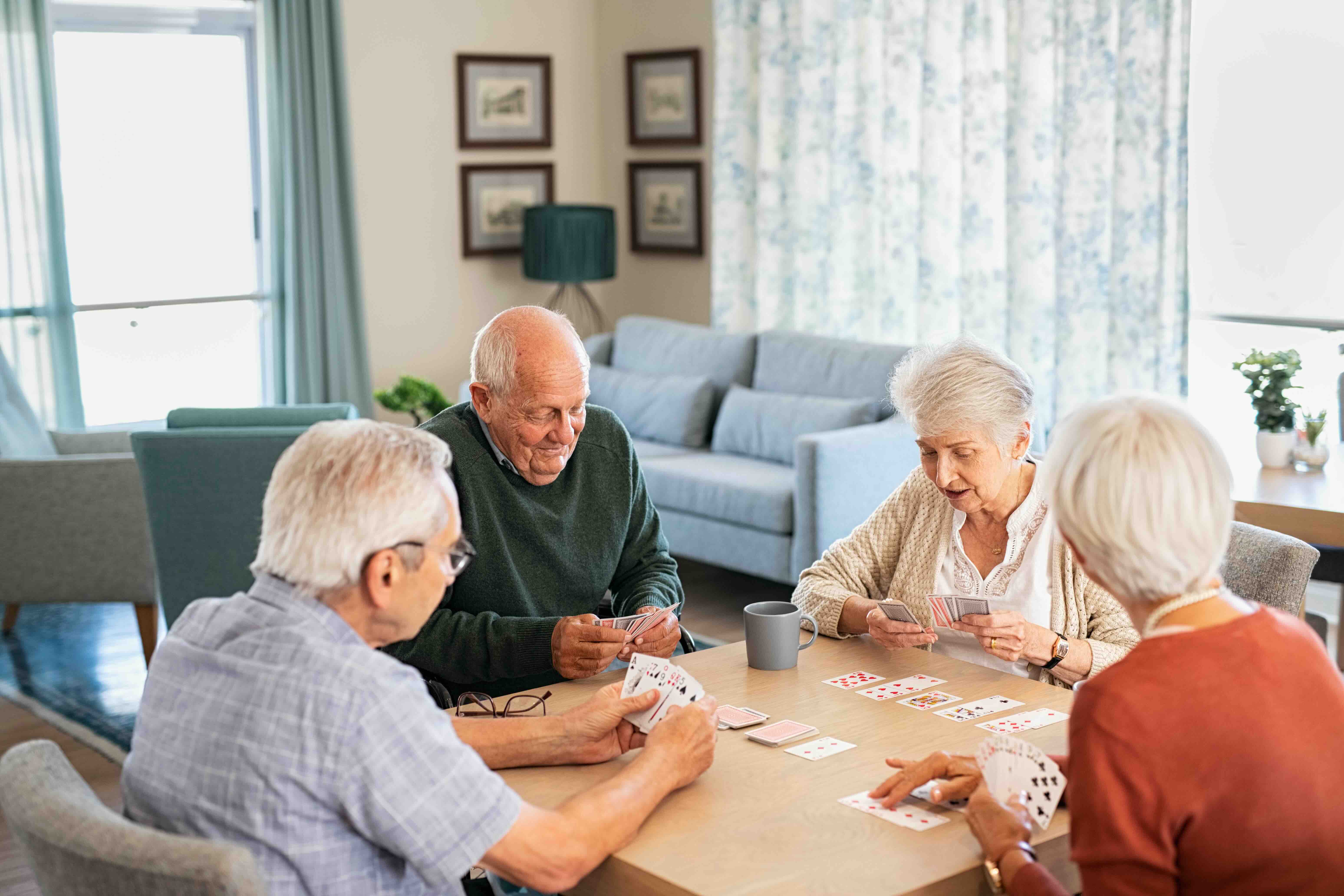 Four older adults smile and play cards together at a dining table in a bright senior living common room.