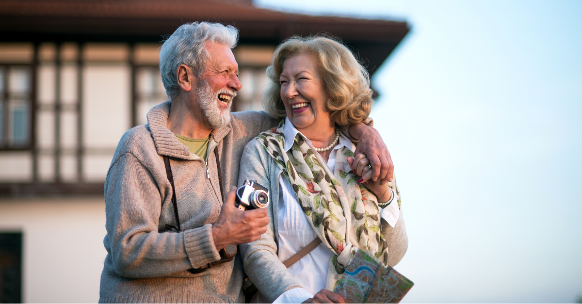 A smiling senior couple laughs together outdoors while sightseeing, the man holding a vintage camera and the woman clutching a travel map, with a charming building in the background.