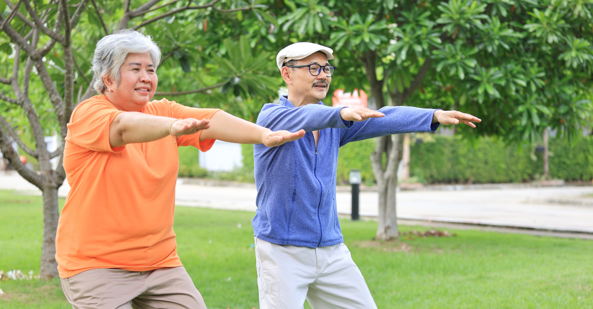 Two older adults practicing tai chi outdoors in a park, smiling and extending their arms in a wide stance, surrounded by lush green trees.