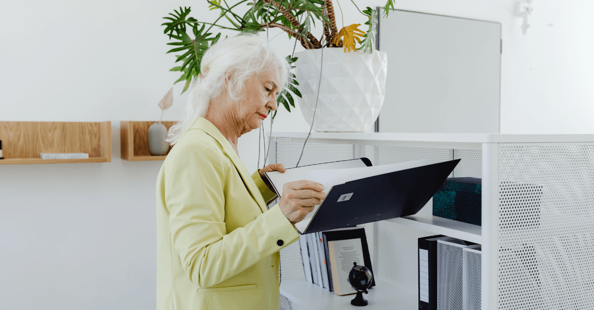 An older woman with white hair wearing a yellow blazer reviews documents in a dark binder while standing at a white shelving unit in a bright, modern space — representing the organizational process of planning a move to a senior living community.