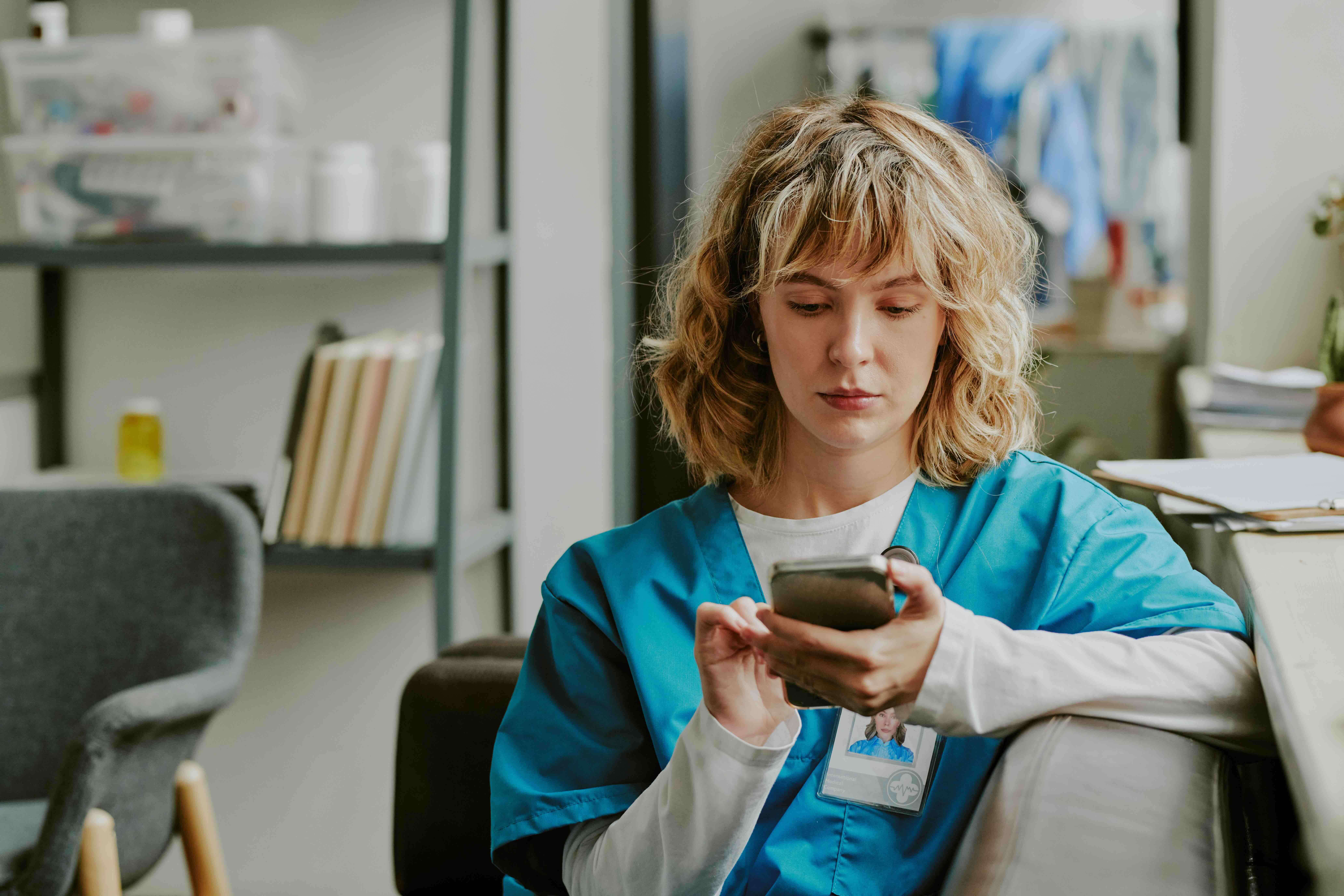A senior living care aide in blue scrubs using a personal cell phone at work, illustrating the HIPAA compliance risk of staff texting resident health information on unsecured personal devices.