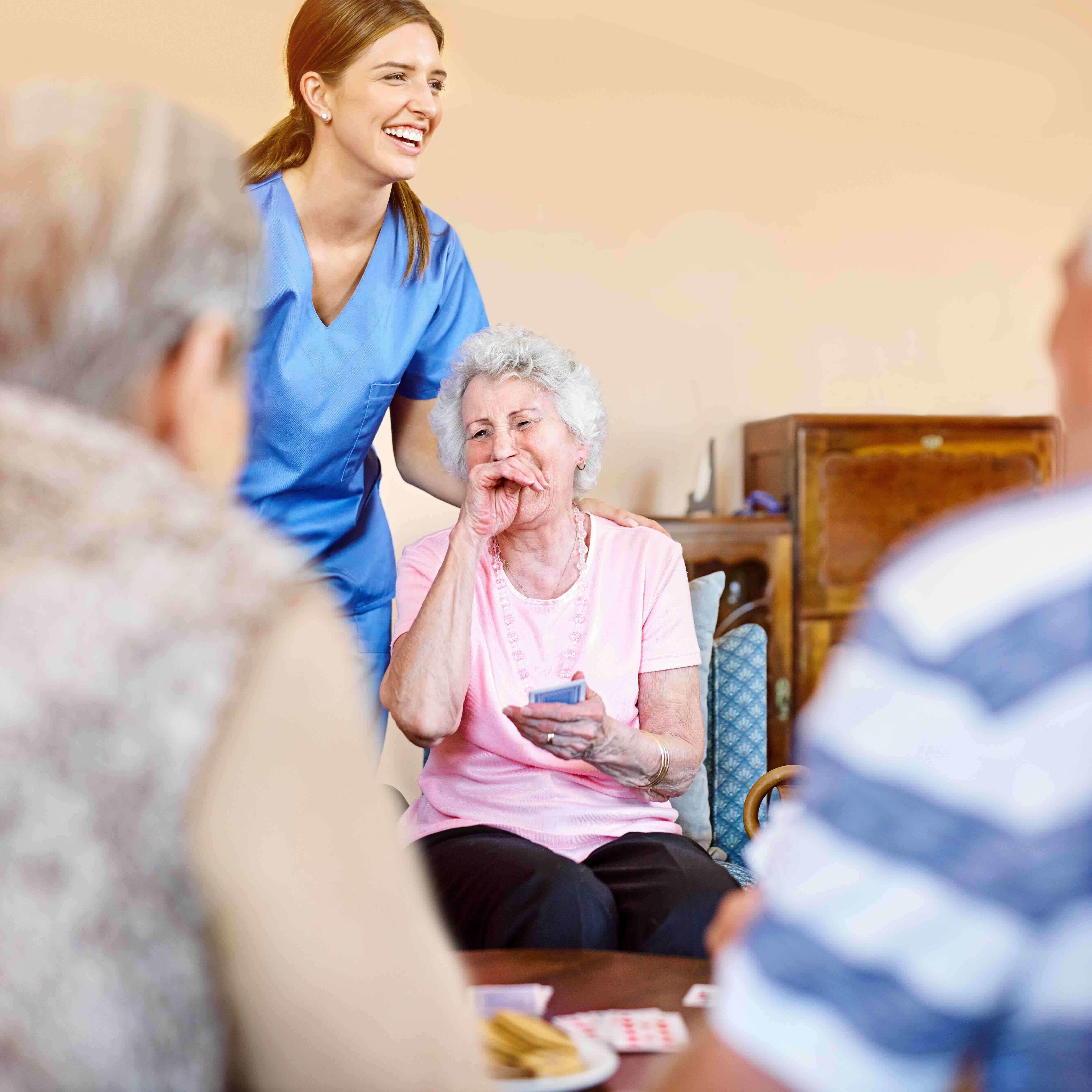 A smiling nurse stands beside an older woman laughing during a card game, reflecting the supportive daily environment that contributes to nursing home costs and the overall cost of nursing home care.