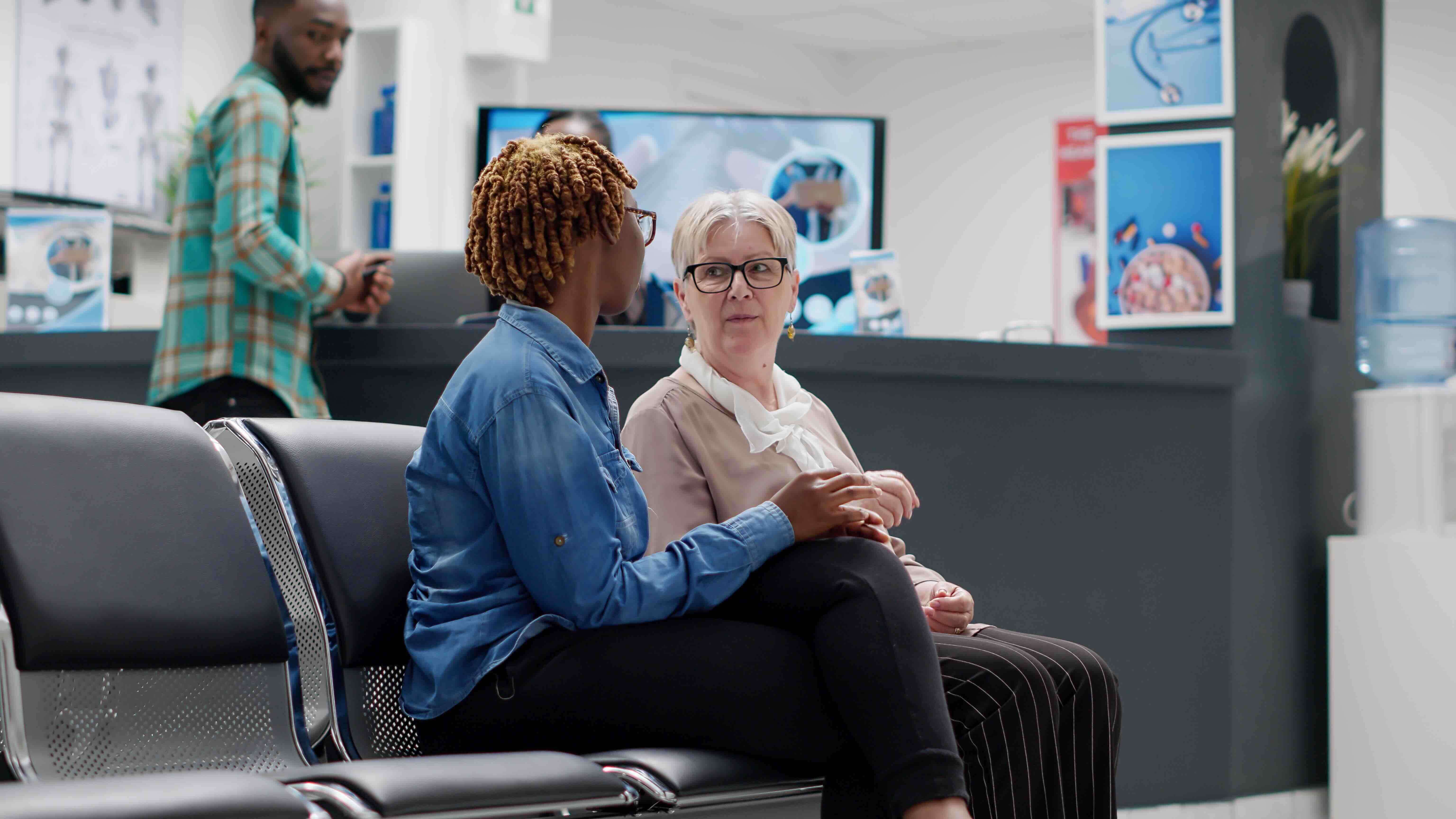 A caregiver sitting beside an older adult in a medical waiting room, offering comfort and support during a stressful moment, visually representing the importance of knowing the emergency action steps, understanding the steps to follow in an emergency are c
